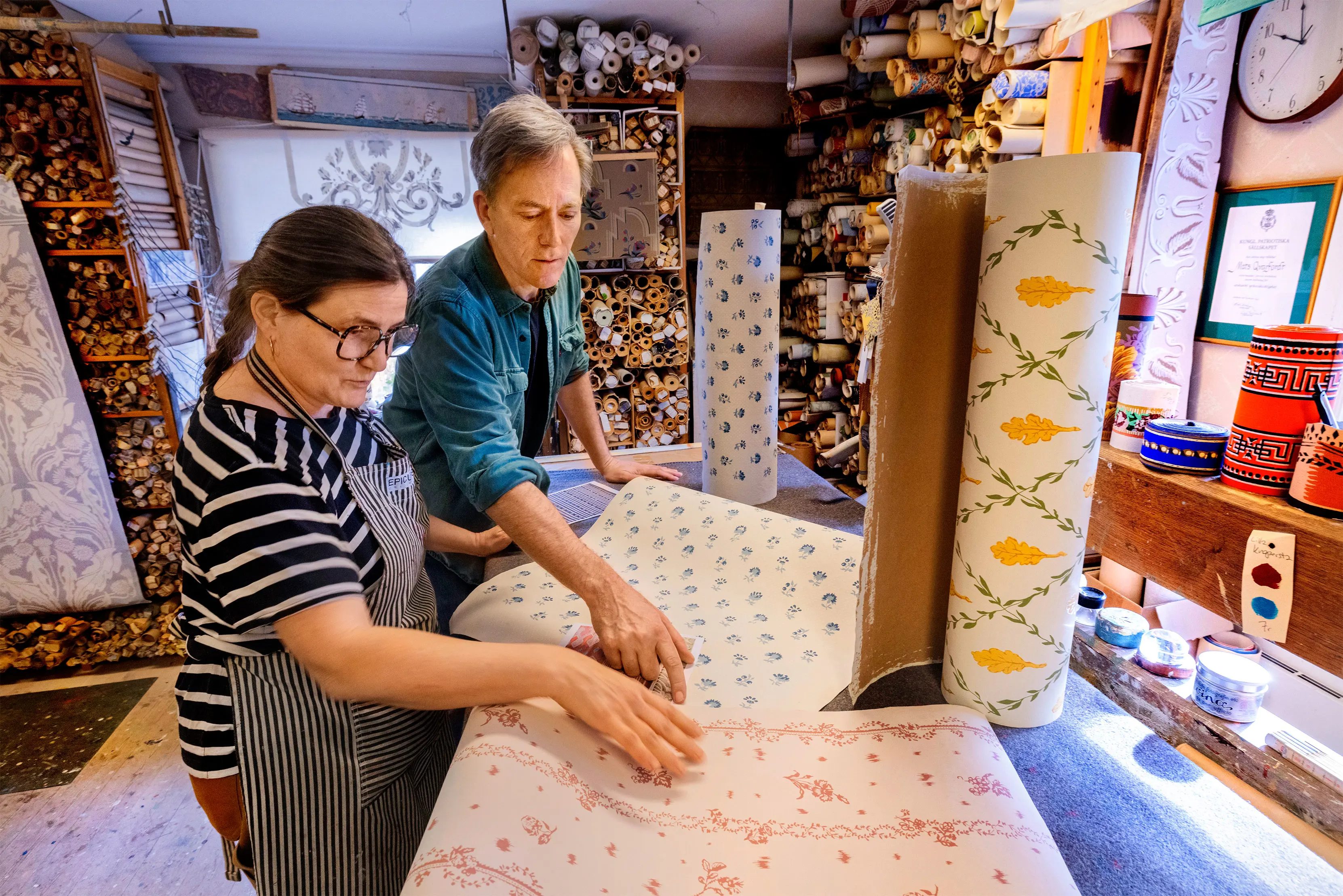 Två personer, man och kvinna, studerar tapetmönster omgivna av tapetrullar  från golv till tak i en verkstad.
