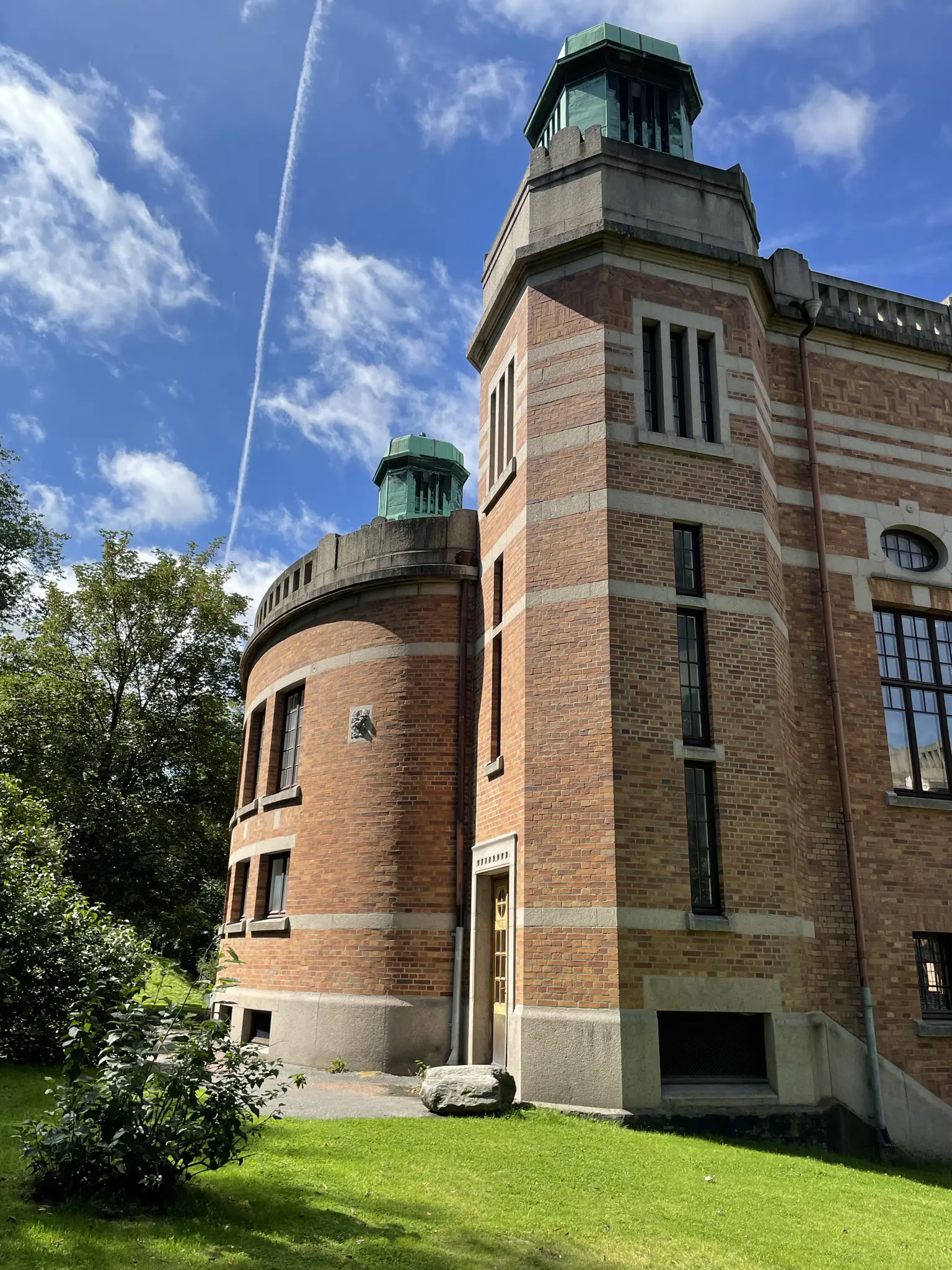 Göteborgs universitet. Massiv byggnad i rött tegel och grå sten. Gaveln har två kopparklädda små torn på taket. Sommar och grönska.