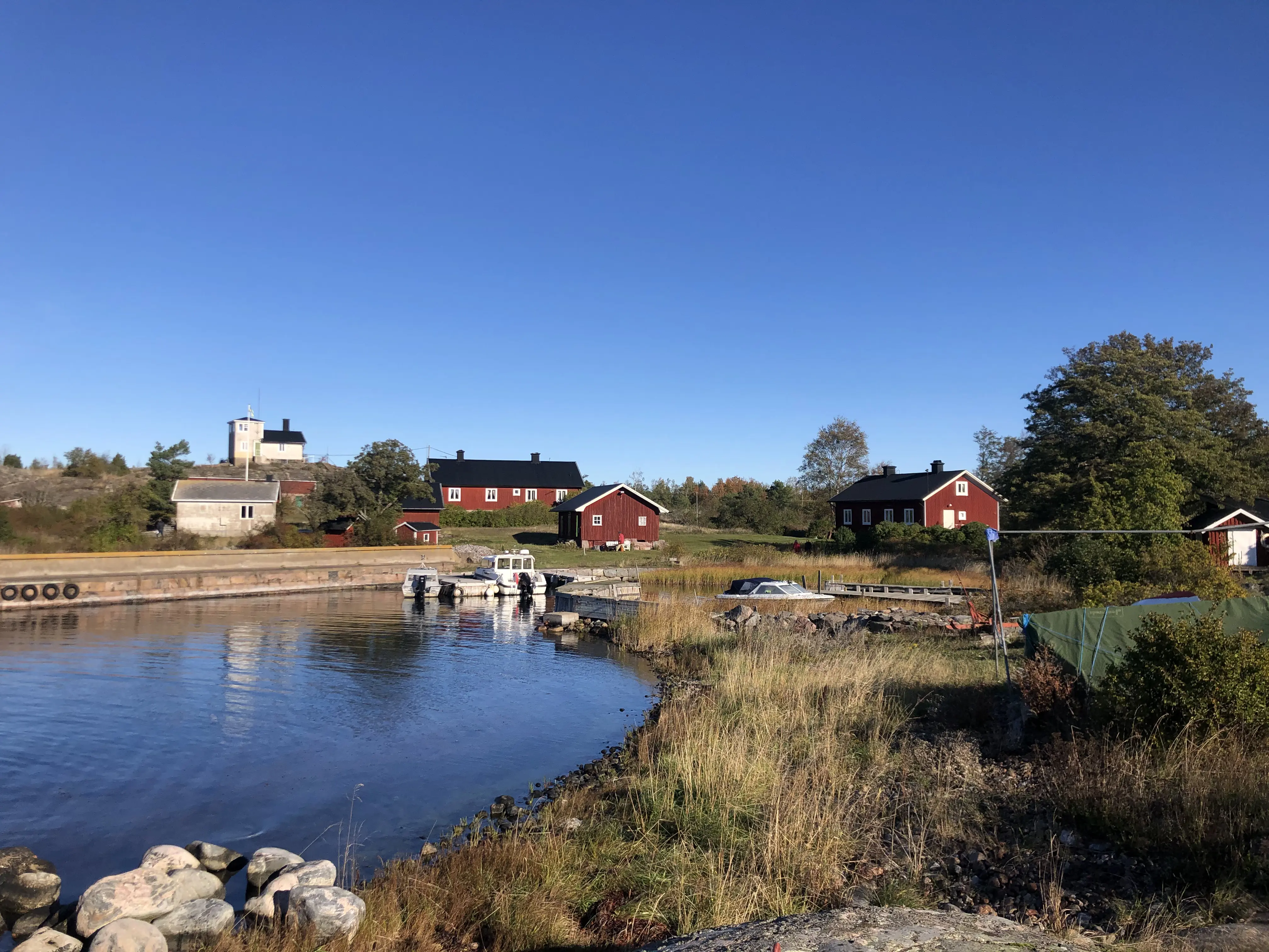 Häradsskär. I en skyddad naturhamn ligger några små båtar. Högt gräs och lummiga träd omger de röda stugorna. På en stenig höjd ligger lotsutkiken.