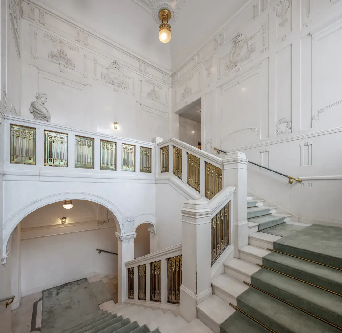 Trapphus med vita väggar, utsmyckade med bland annat Sveriges riksvapen. I trappan ligger en grön matta.