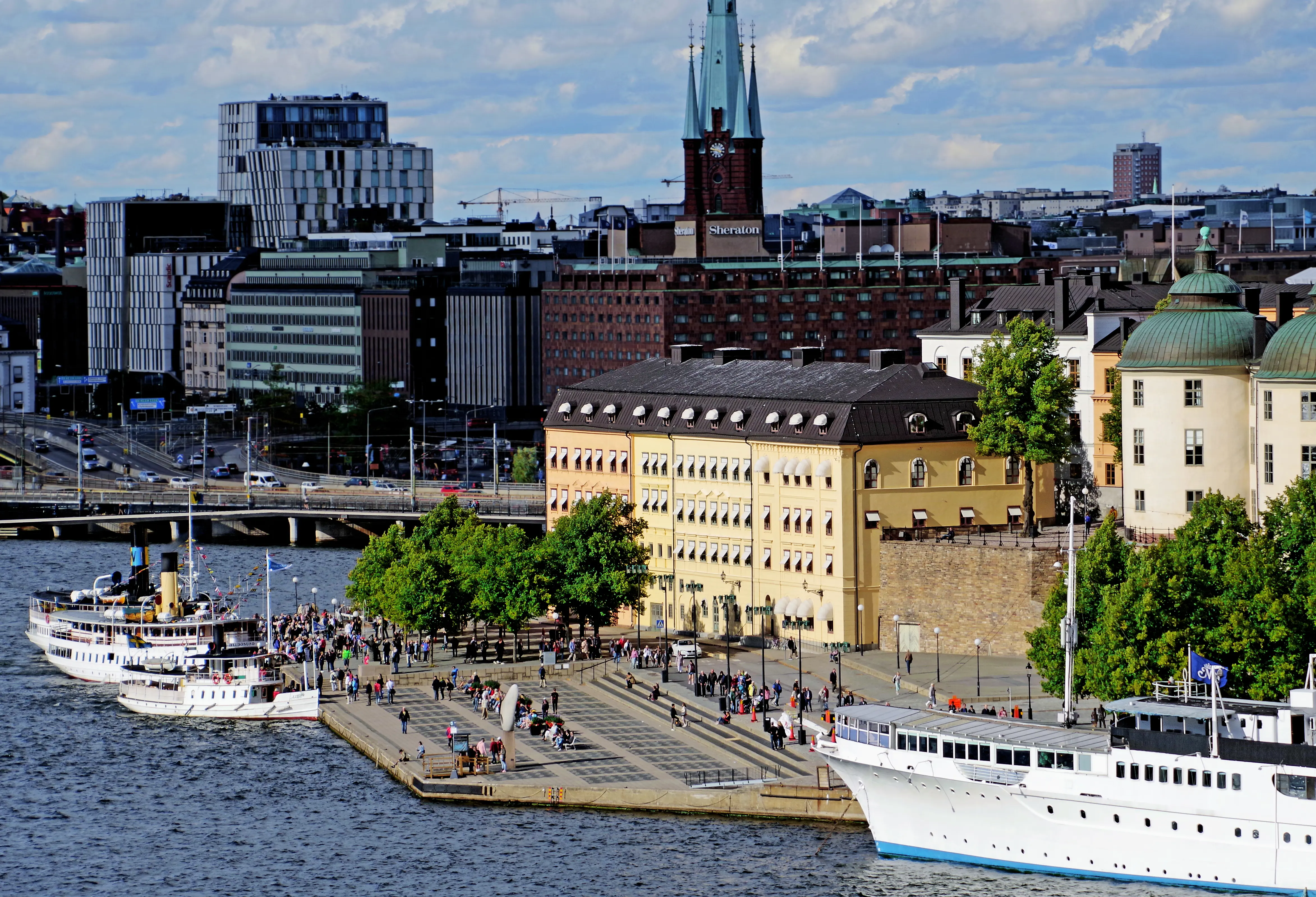 Riddarholmen sedd från Södermalm. På västra kajen promenerar många människor. I vattnet ligger en rad ångbåtar.