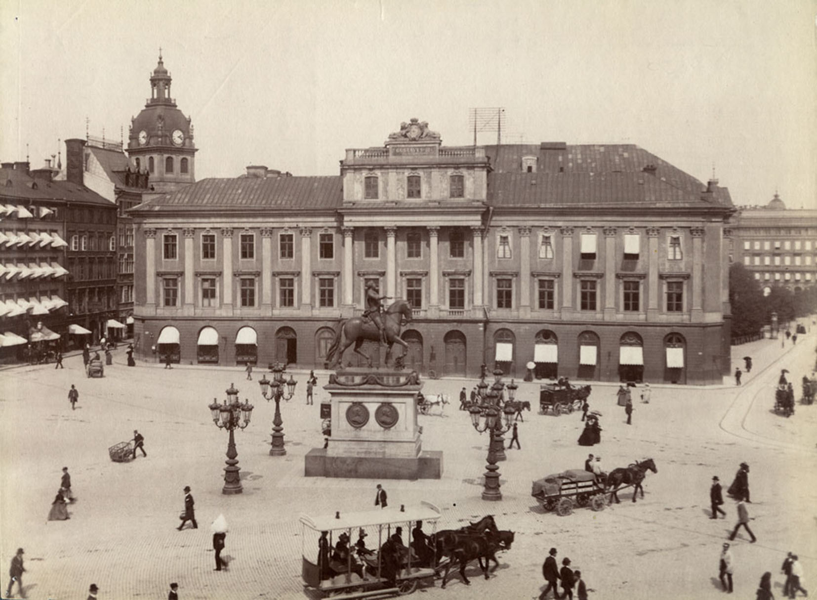 Svartvit bild av stort hus vid ett torg. På torget är människor och en häst-spårvagn.