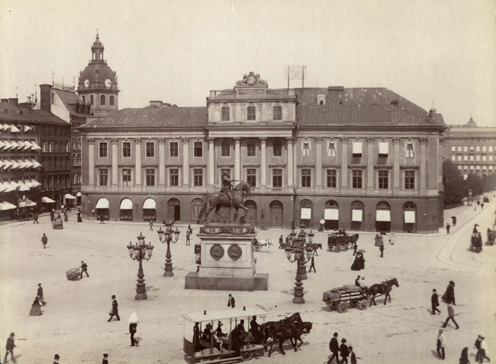Svartvit bild av stort hus vid ett torg. På torget är människor och en häst-spårvagn.