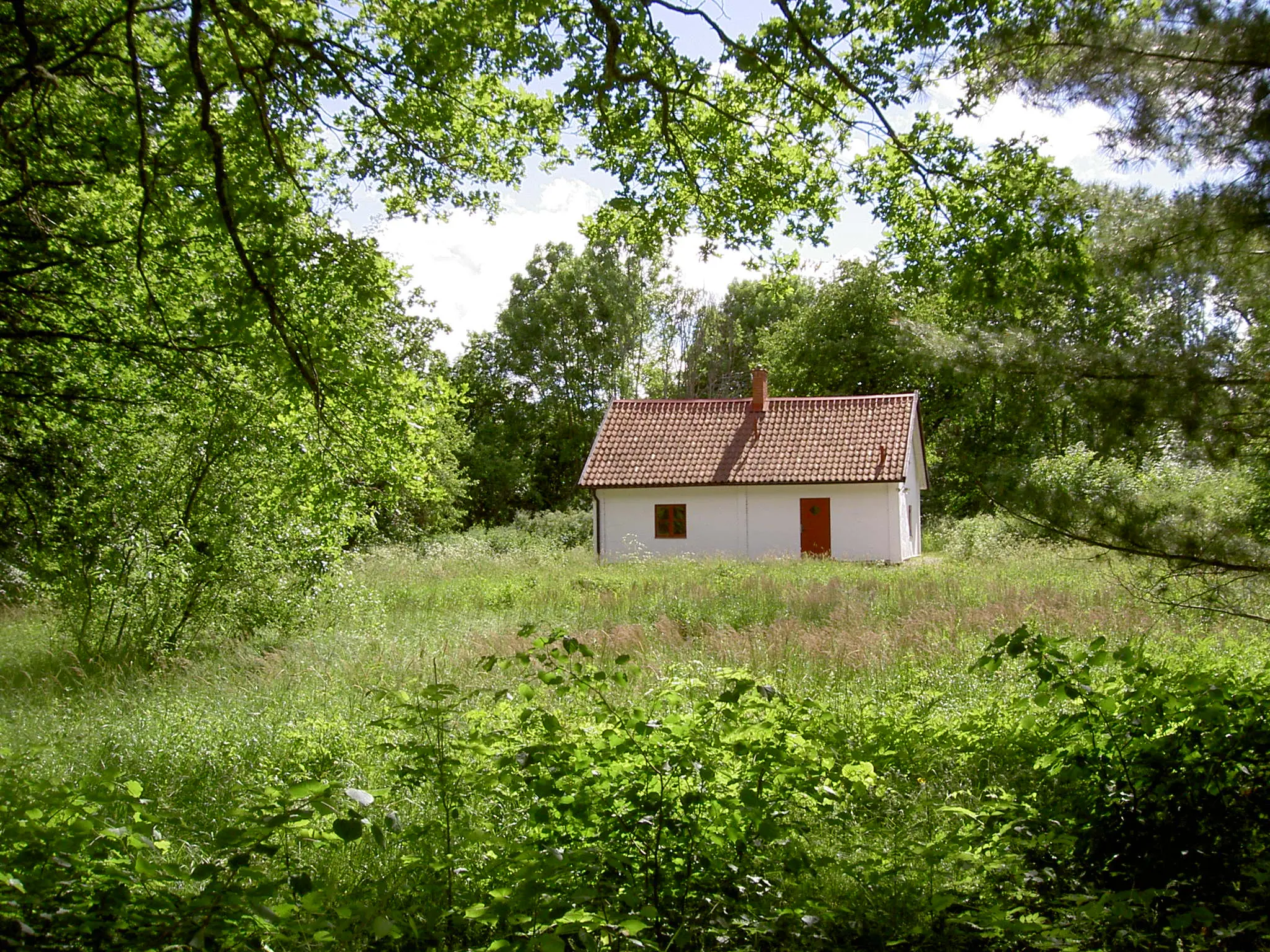 Trädgårdsmästarbostaden på Åholmen. Ett vitputsat litet hus med rött tegeltak, omgivet av blommande gräsäng och lummiga träd.
