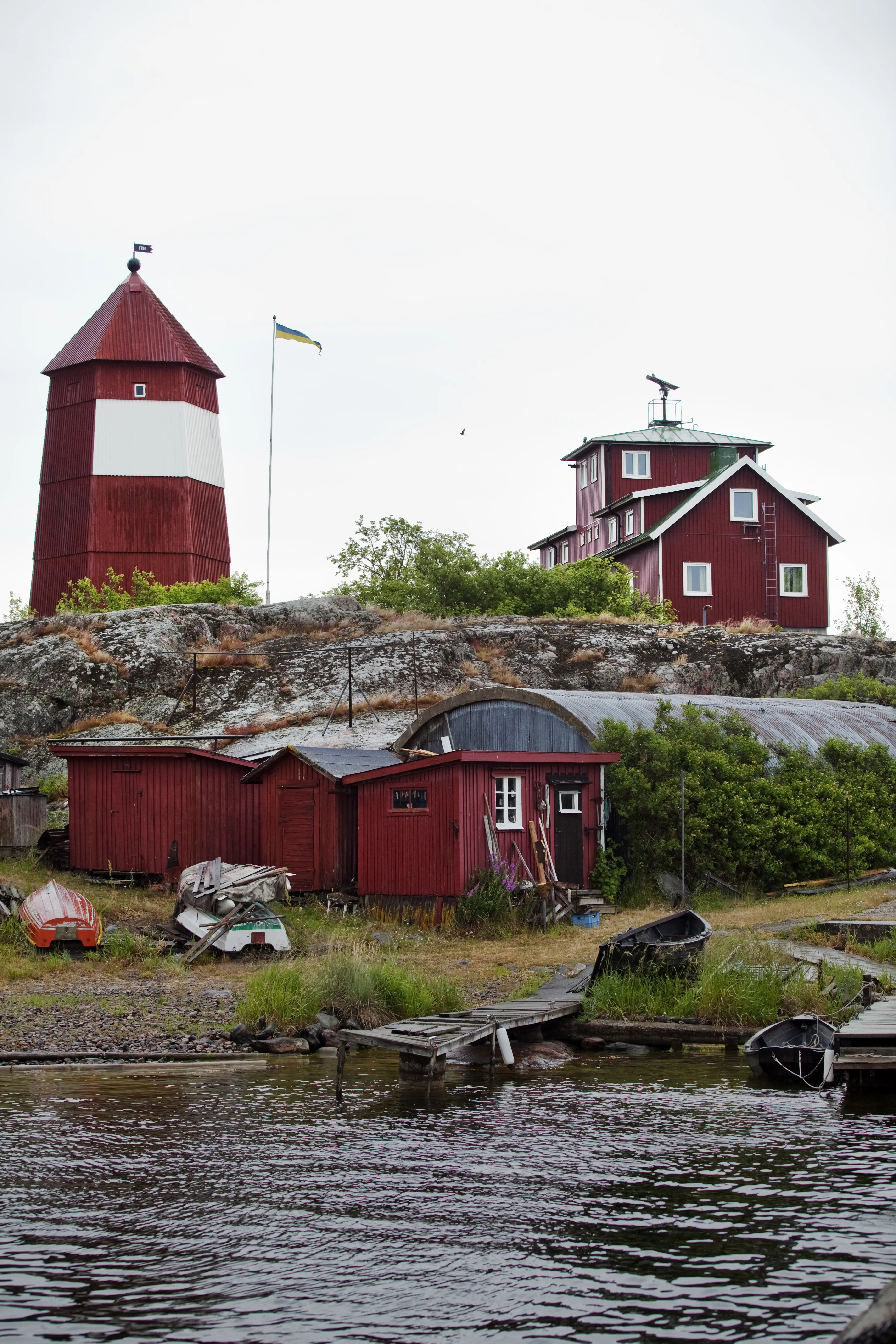 Hävringe båk, litet fyrtorn i rödmålat trä med vitt parti på mitten. Vid vattnet ligger små bodar, bryggor och ekor.
