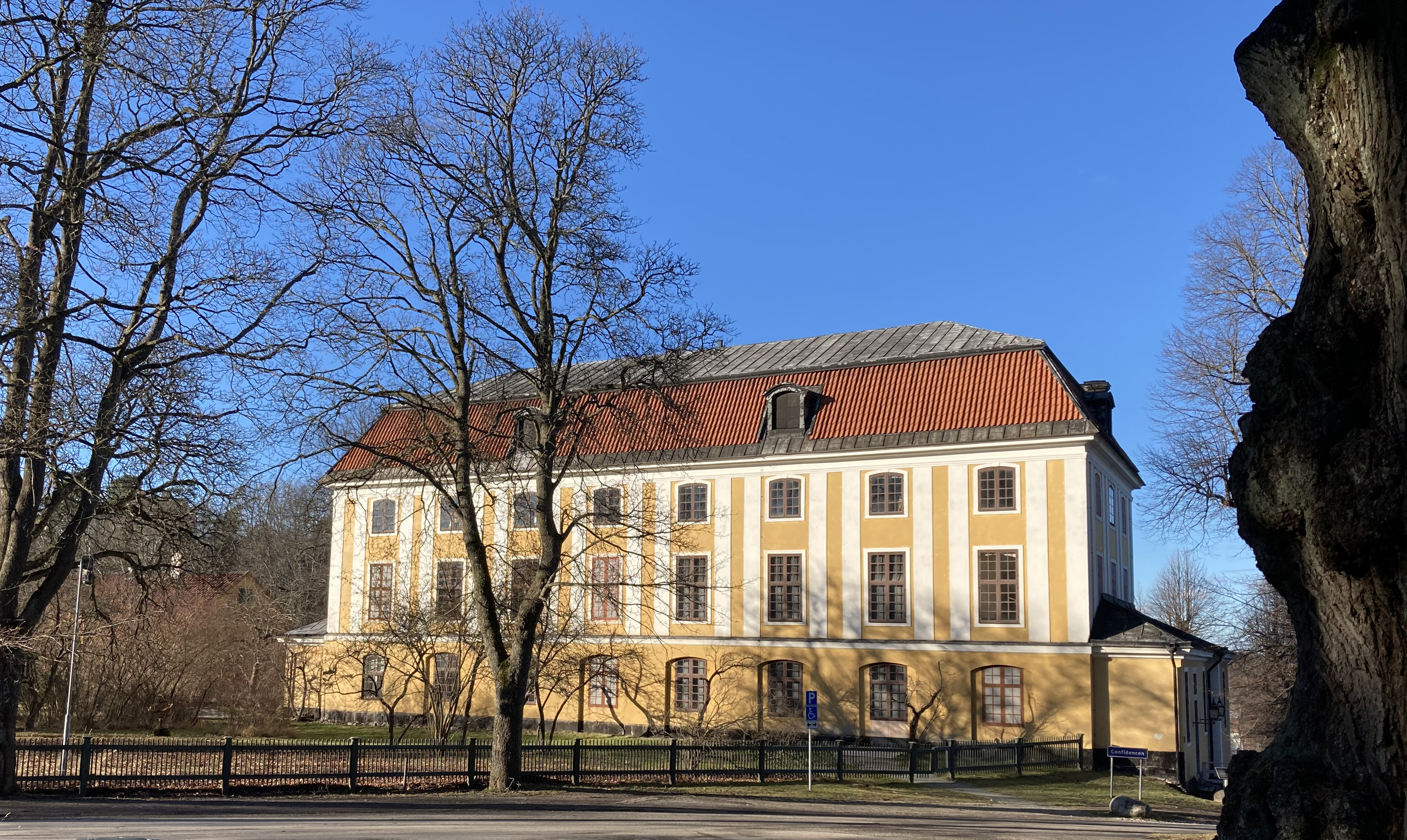 Confidencen, slottsteatern vid Ulriksdals slott. Byggnad i tre våningar med gul och vit puts. Tidig vår, träden står ännu nakna.
