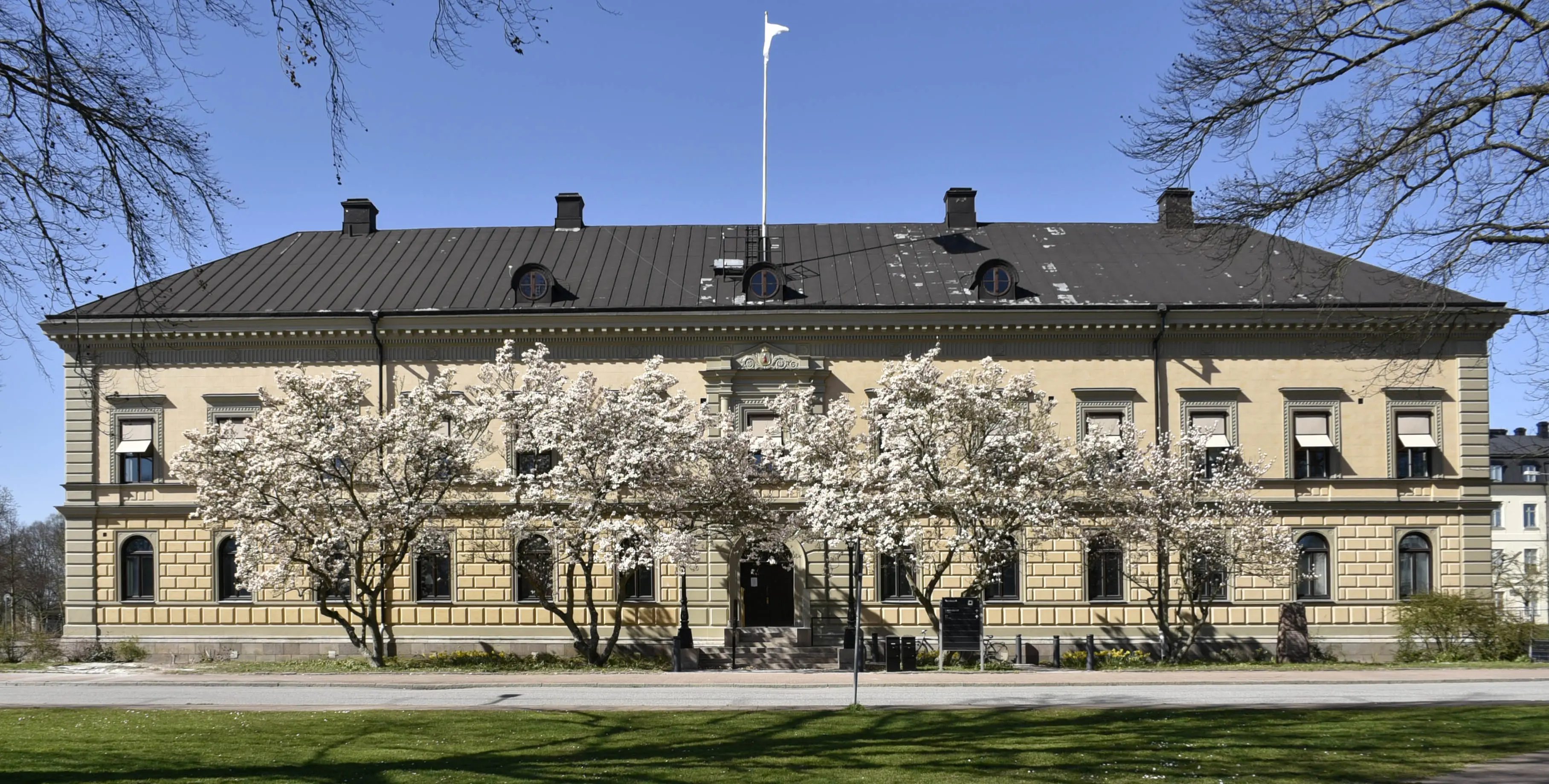 Kristianstads residens. En beige-putsad tvåvåningsbyggnad med svart tak. Framför porten blommar körsbärsträd.