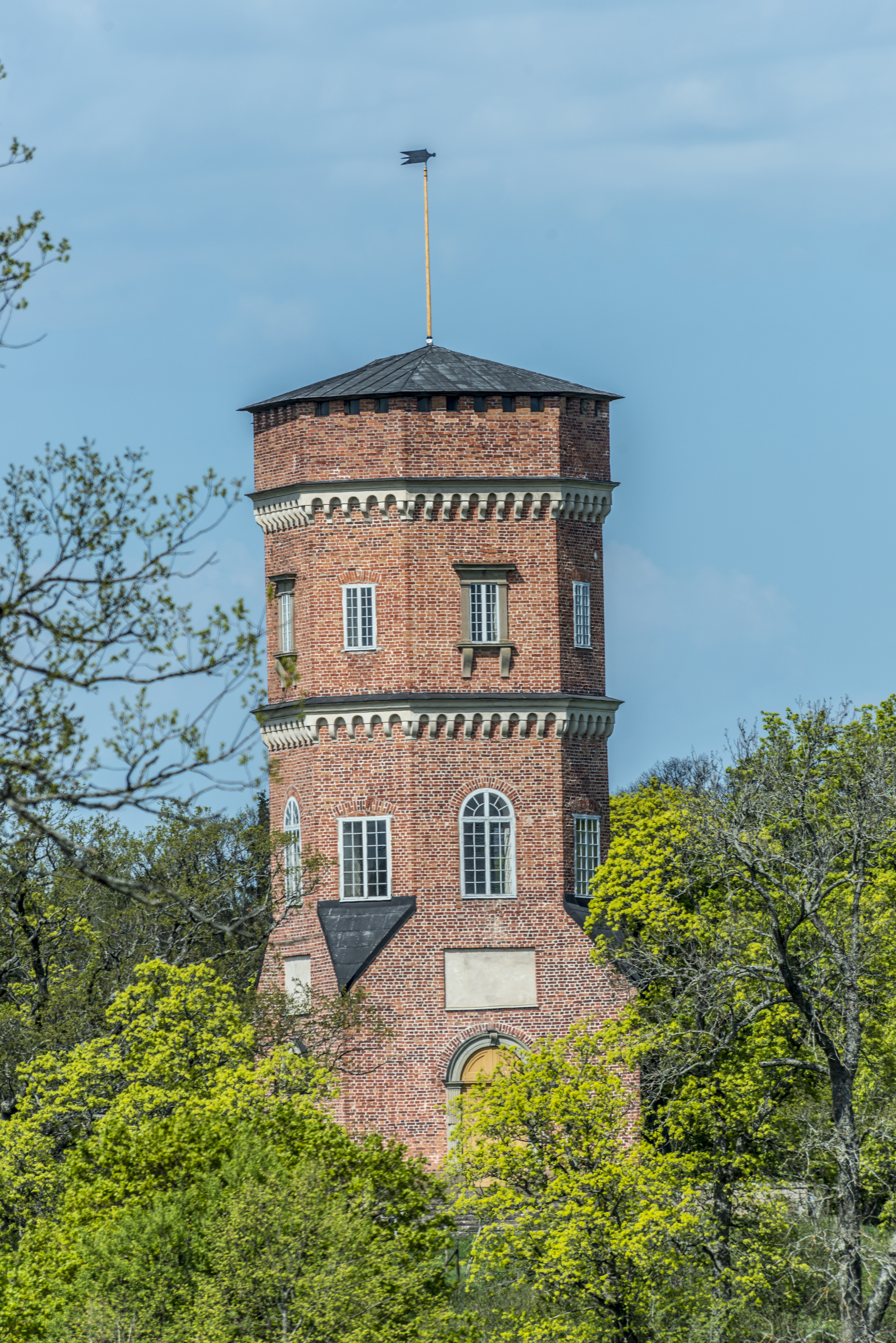 Götiska tornet vid Drottningholm. Åttakantig tegelbyggnad på en liten höjd, omgiven av träd. Vår och grönska.
