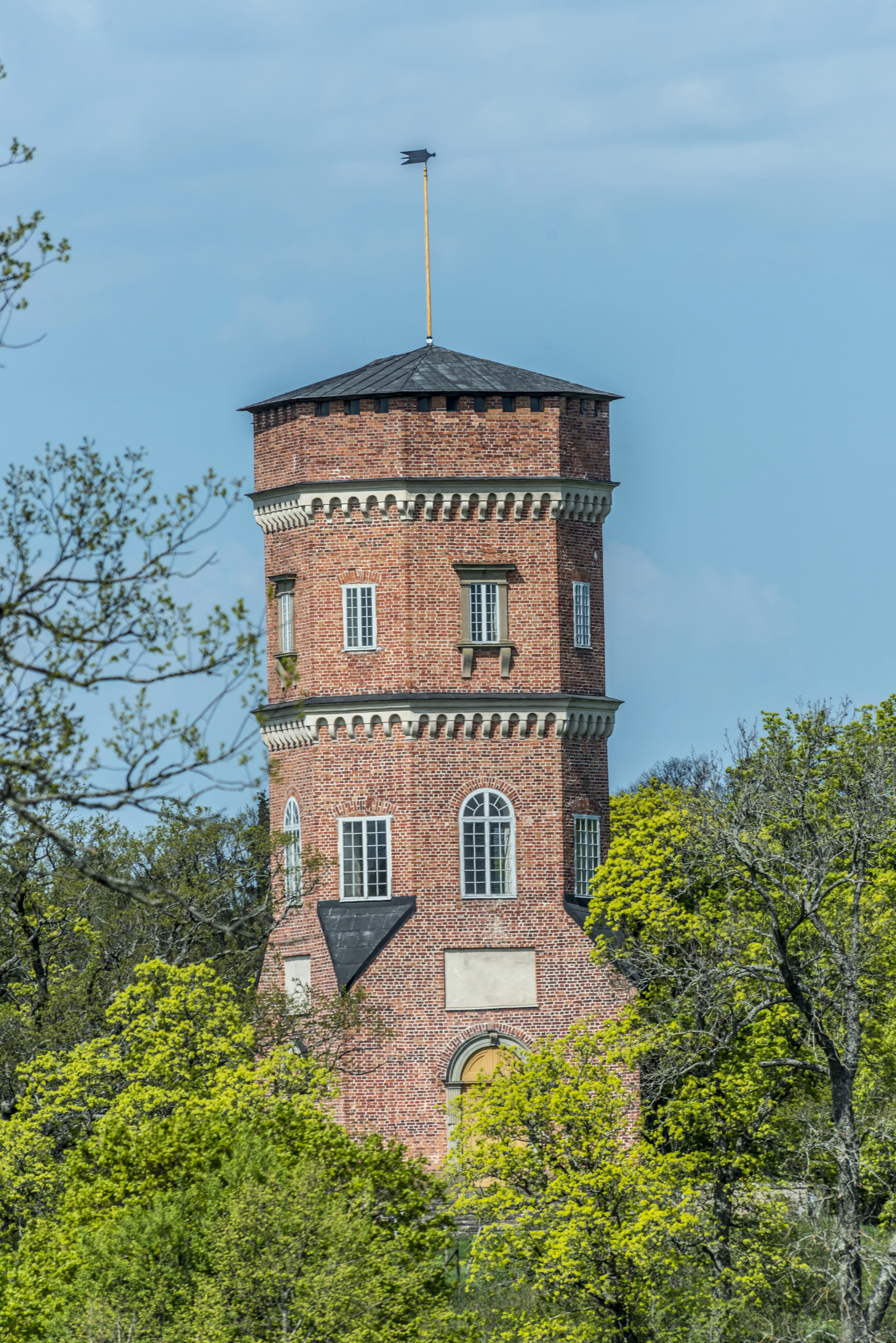 Götiska tornet vid Drottningholm. Åttakantig tegelbyggnad på en liten höjd, omgiven av träd. Vår och grönska.