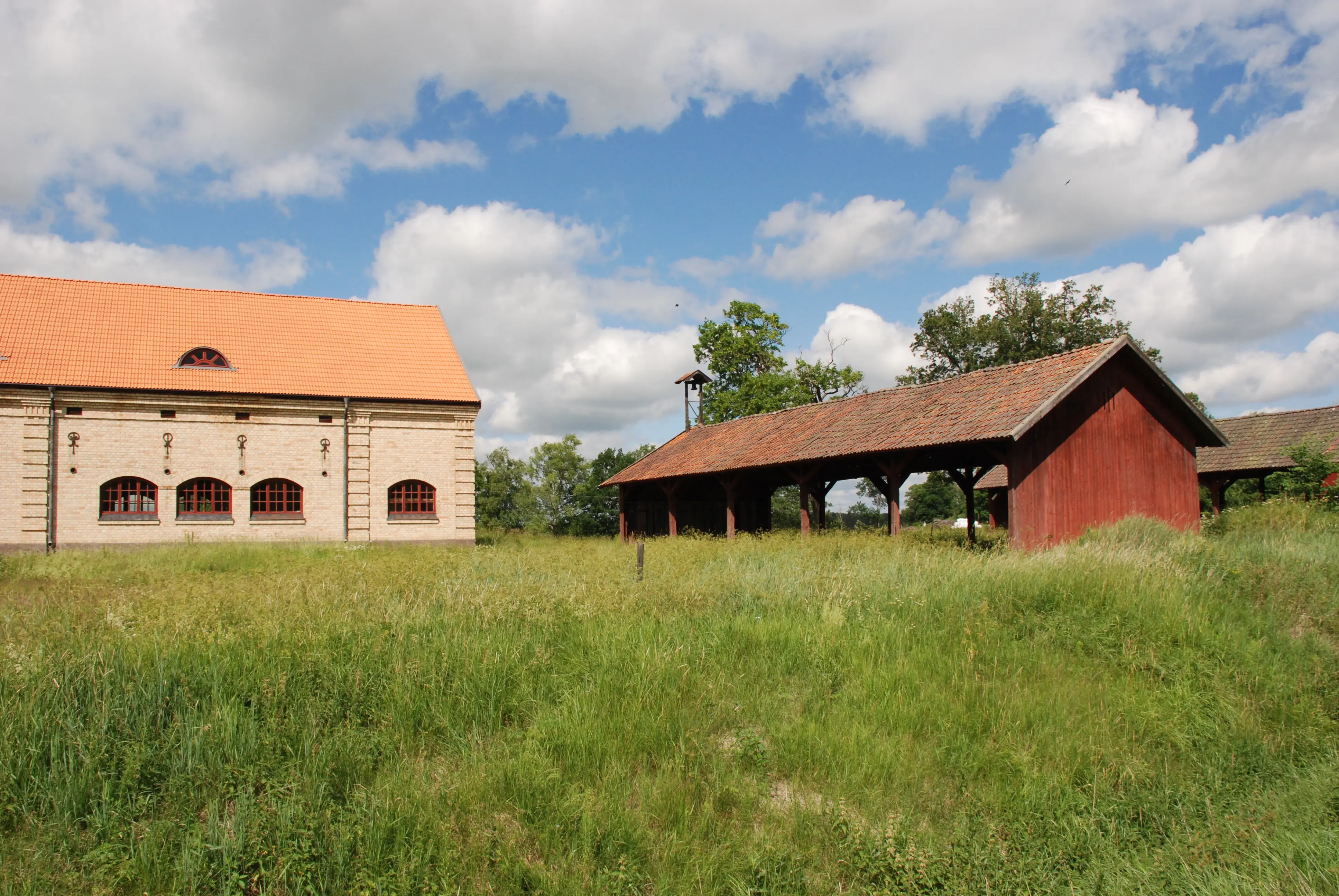 Salsta kronoegendom, ekonomibyggnader. Ett stort stall byggt i sten och ett öppet vagnslider i rödmålat trä. Sommar och grönska.