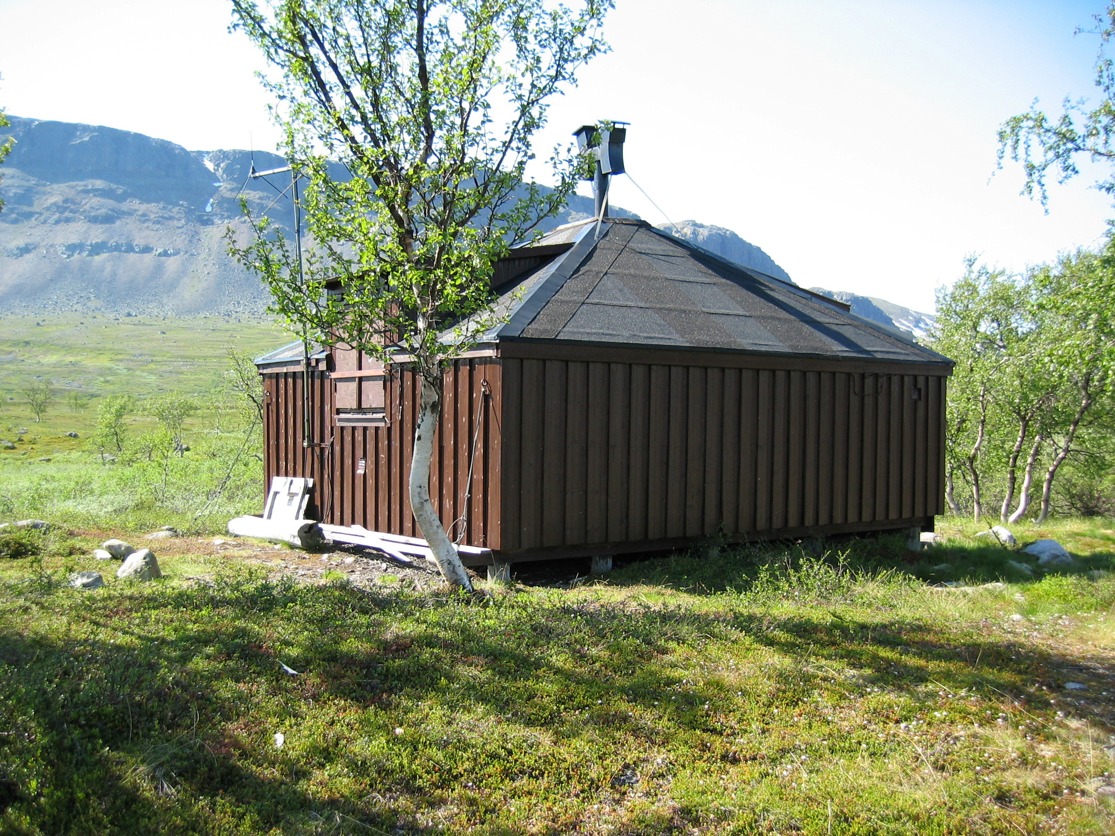 Kvadratisk trästuga utan fönster, med tak av tjärpapp. Tundra med enstaka fjällbjörkar. Ett berg med snöfläckar.
