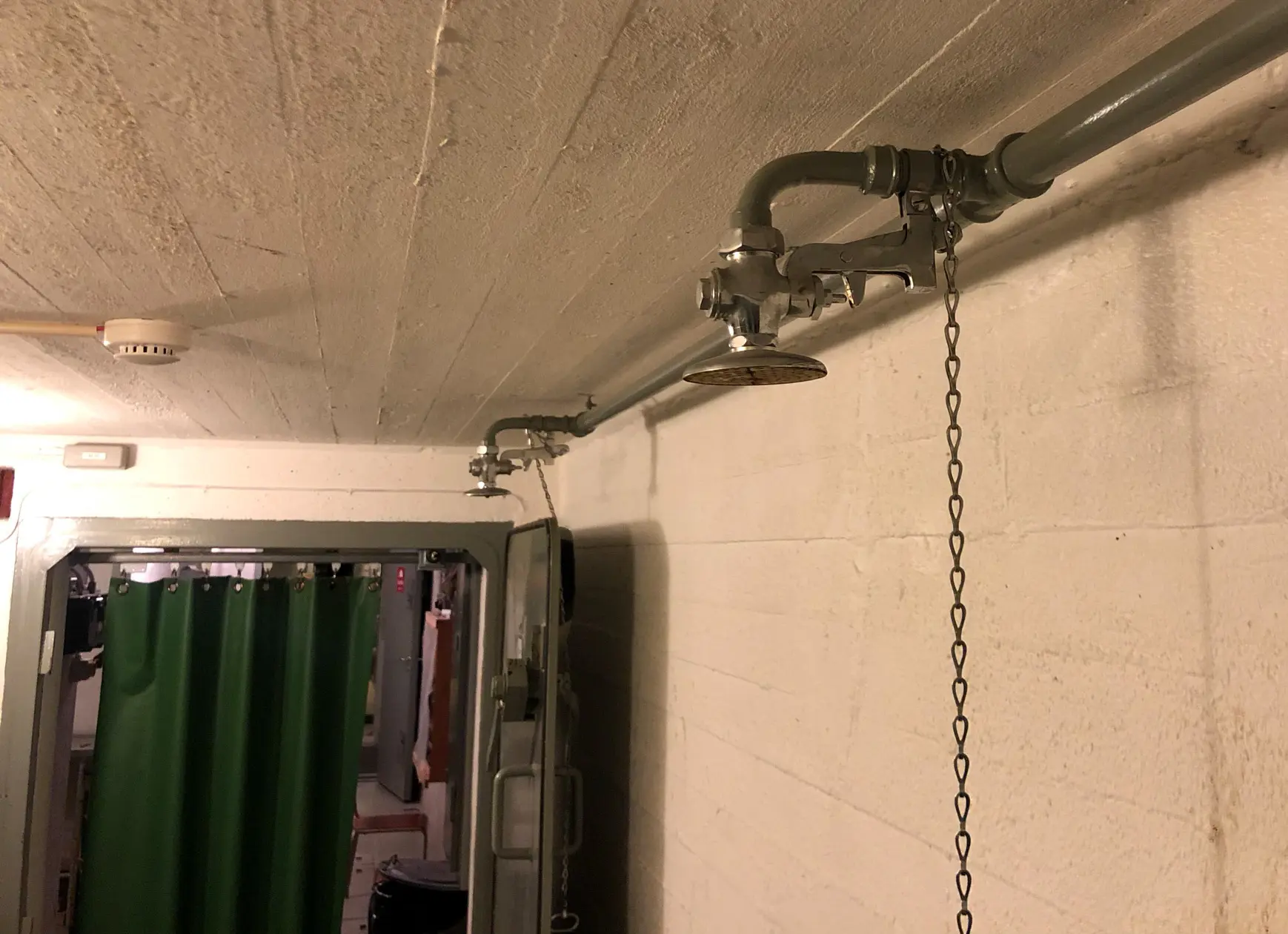 Ett genomgångrum med vitmålade betongväggar. Två duschmunstycken av metall fästa på en vattenledning vid taket och framför en dörr anas ett tjockt grönt gummidraperi.
