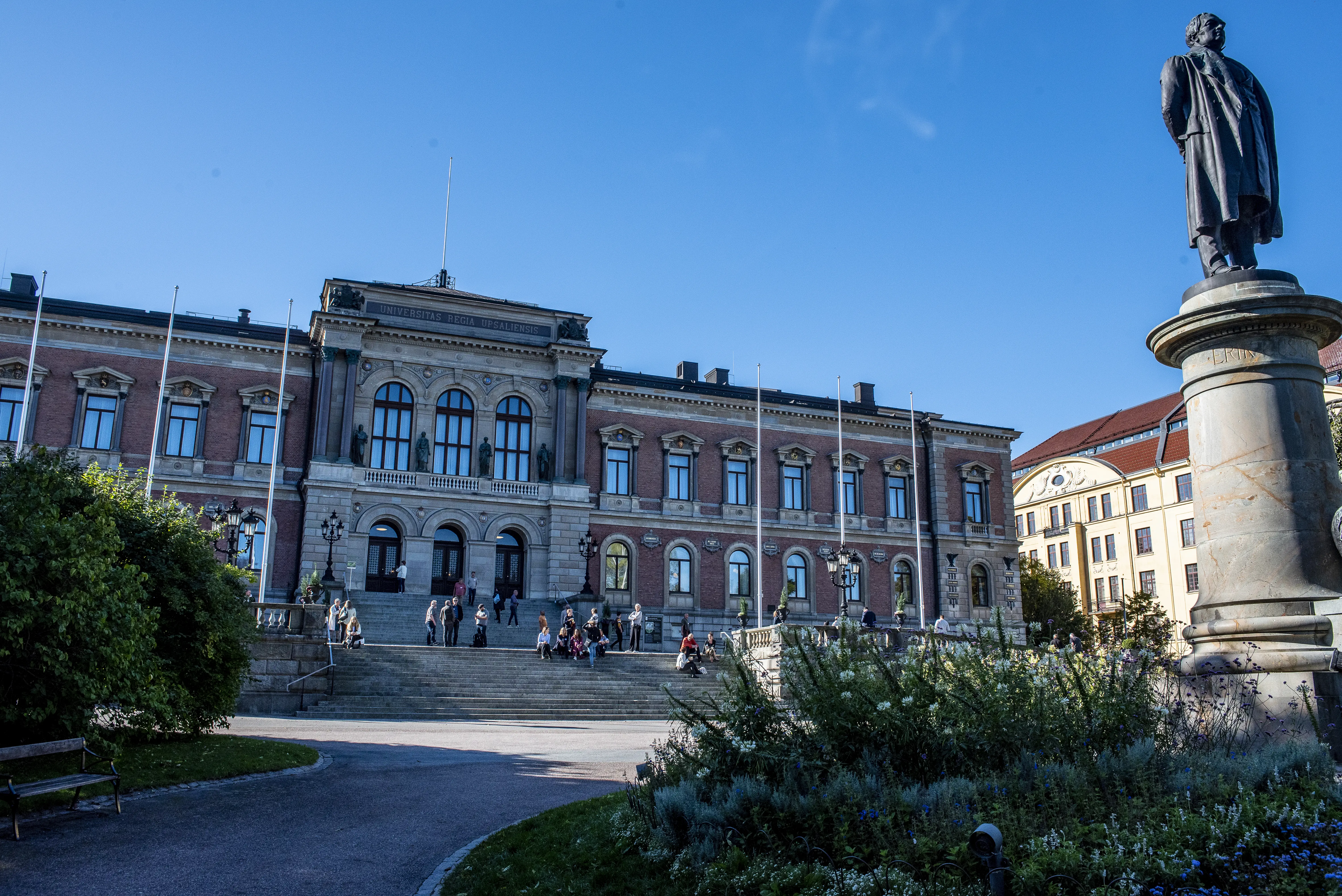 Uppsala universitetshus. Tvåvåningsbyggnad klädd i rött tegel, med klassicistisk stendekor. På trapporna till entrén sitter och står ett antal personer.