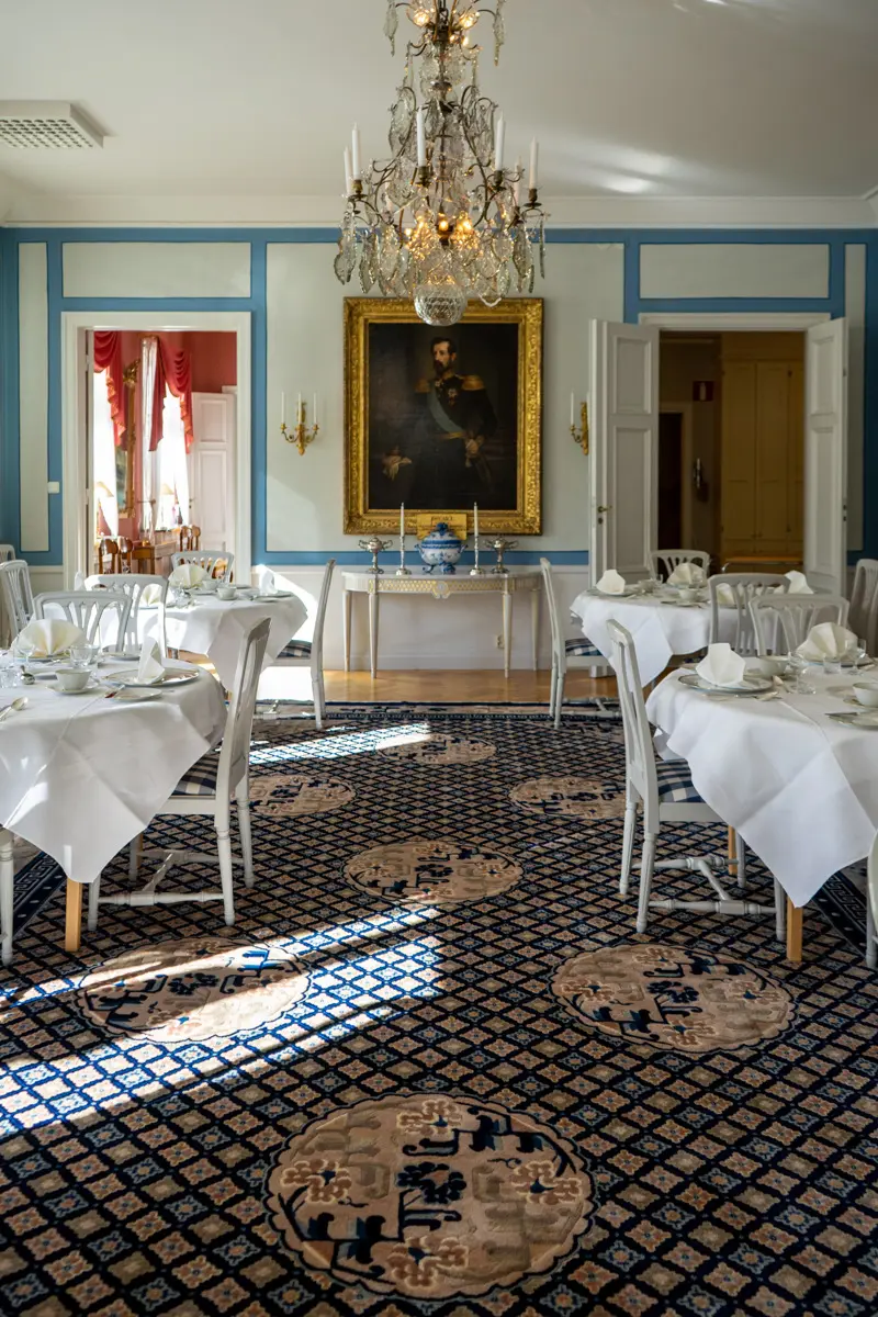 Matsal med ett porträtt i guldram, kristallkrona och bord med vita dukar.