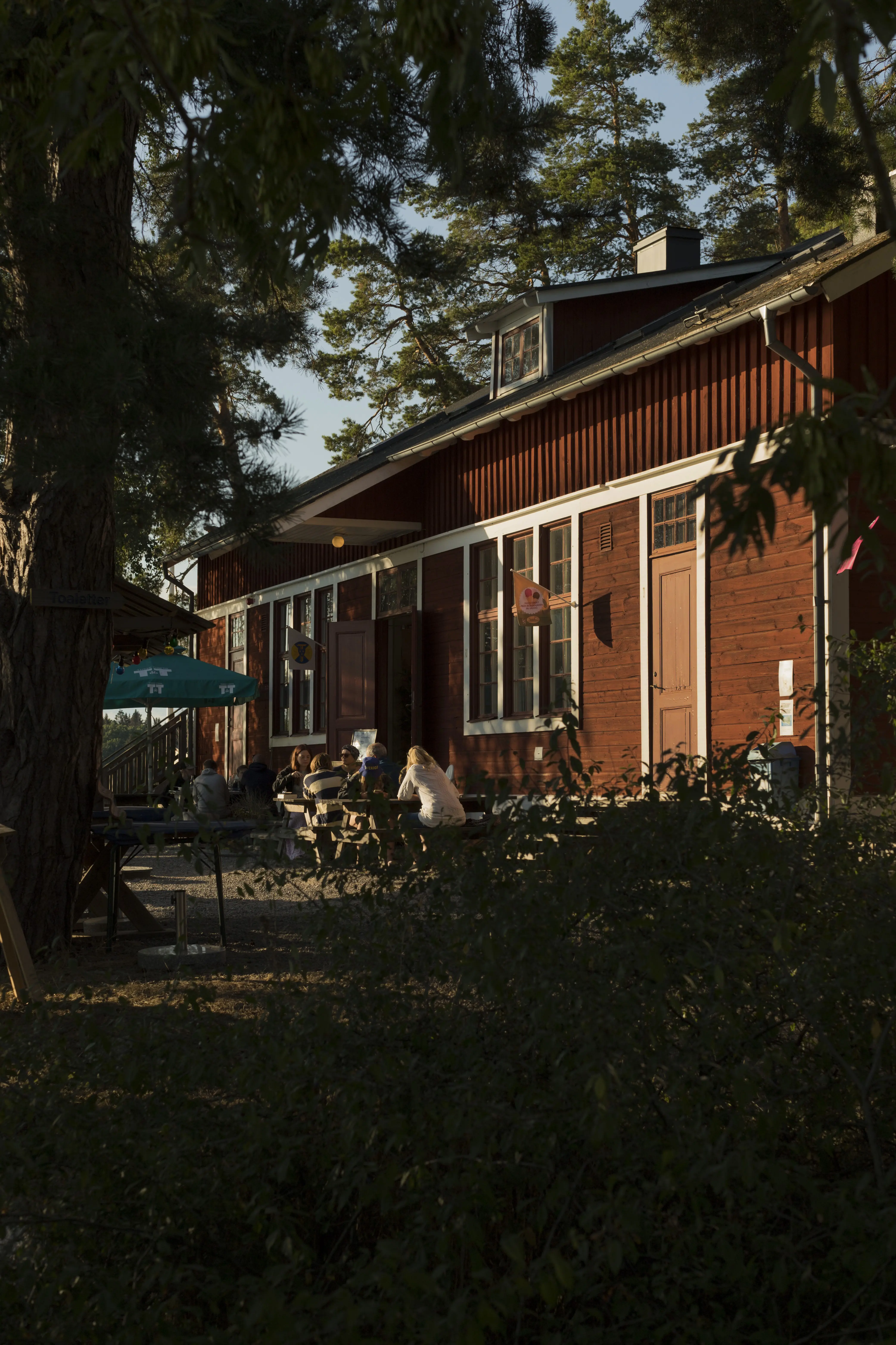 Siarö fort. Besökare på uteserveringen vid det röda trähuset som rymmer kafé och vandrarhem. Sommar och grönska.