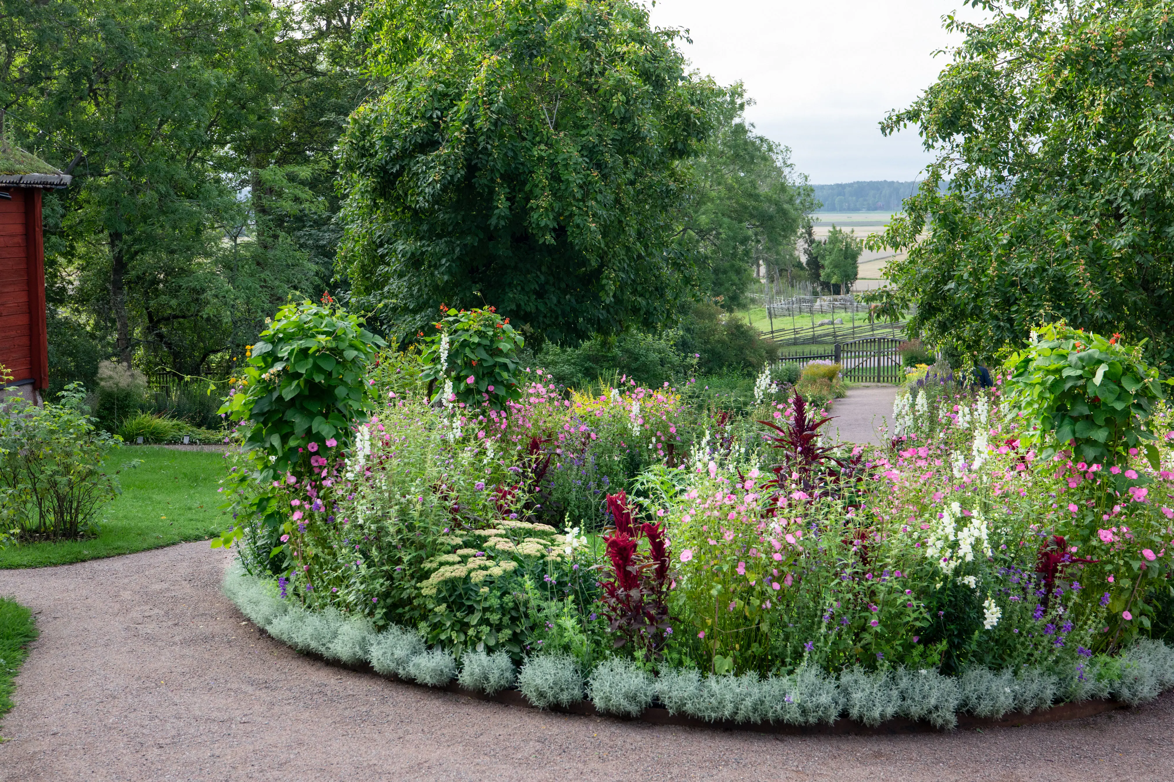 Rund rabatt med blommor, omgiven av grusgång, med landsbygdslandskap i bakgrunden.