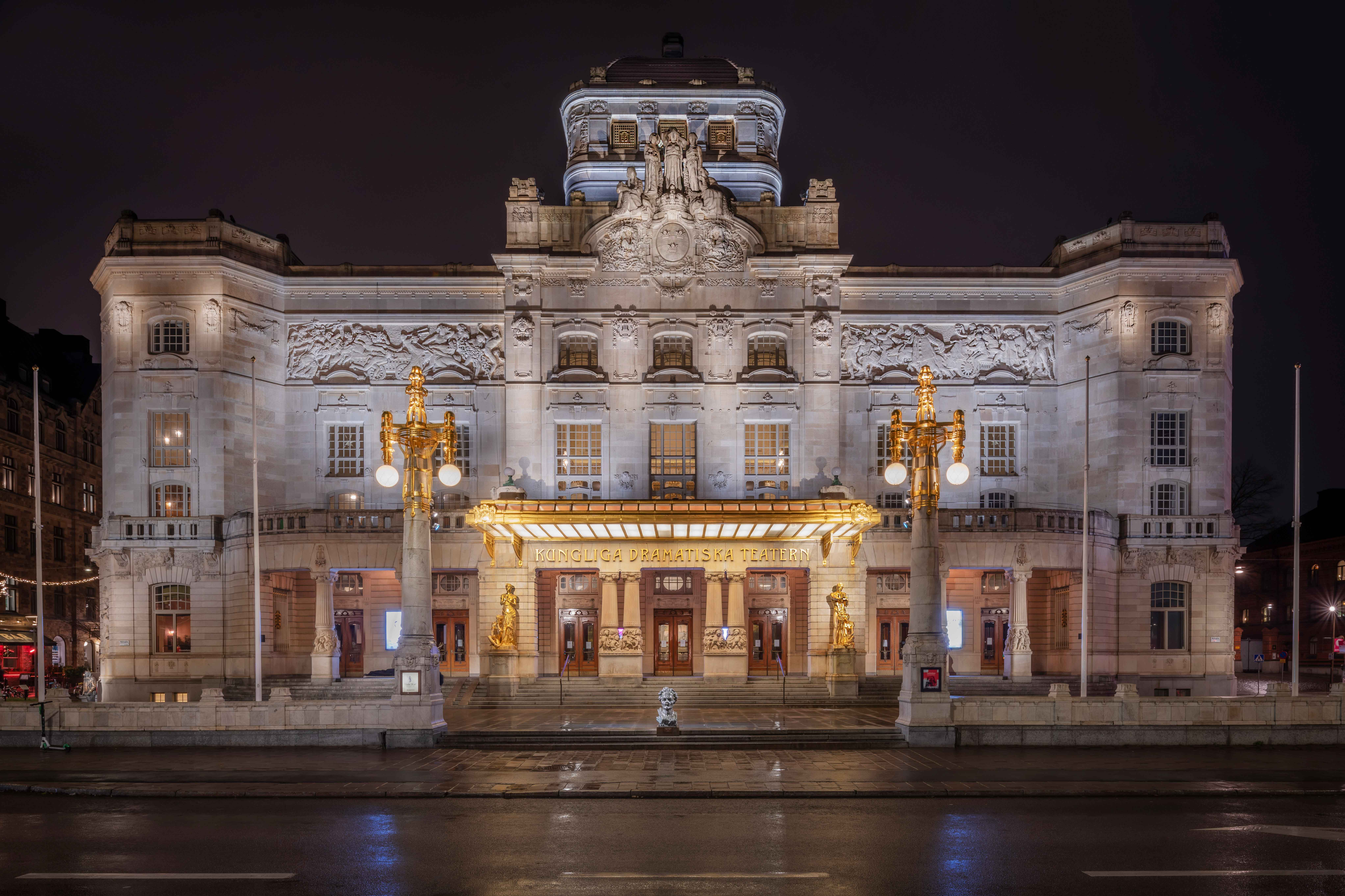 Dramaten sedd från huvudentrén mot mörk kvällshimmel och upplyst med fasadbelysning.