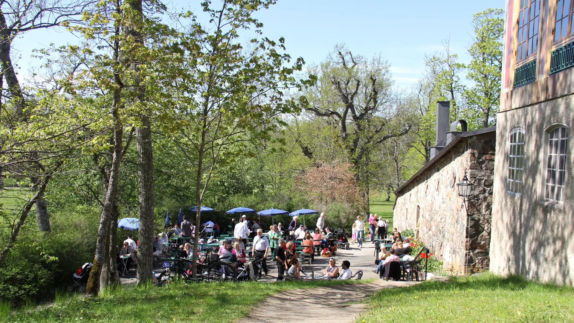 Slottsköket vid Kina slott, Drottningholm. En låg stenbyggnad. På en uteservering sitter besökare. Vår och grönska.