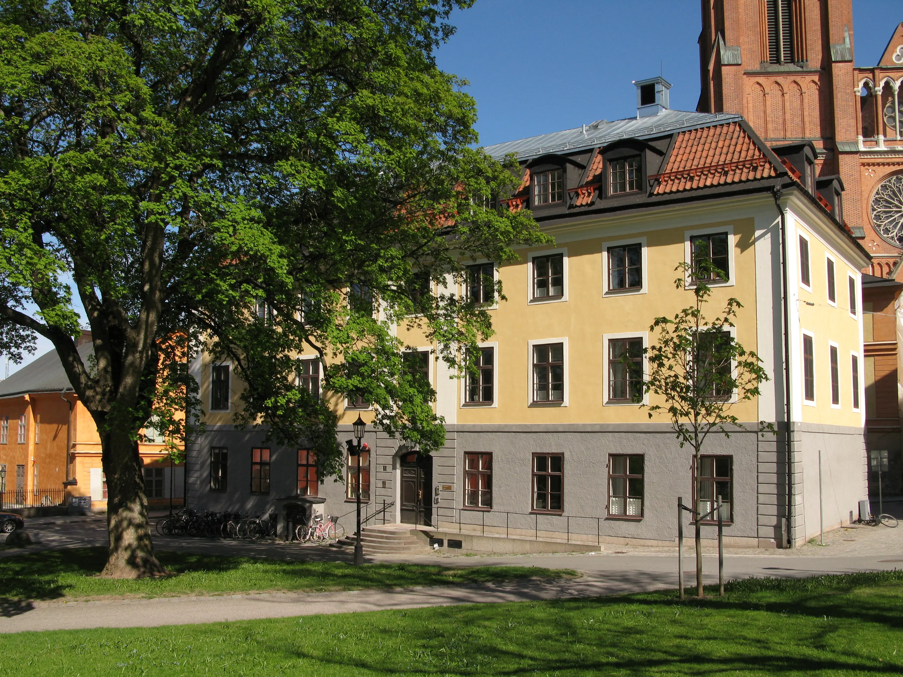 Ekermanska huset i Uppsala. Ett gulputsat trevåningshus med tak-kupor. Alldeles intill ligger domkyrkan. Sommar och grönska.