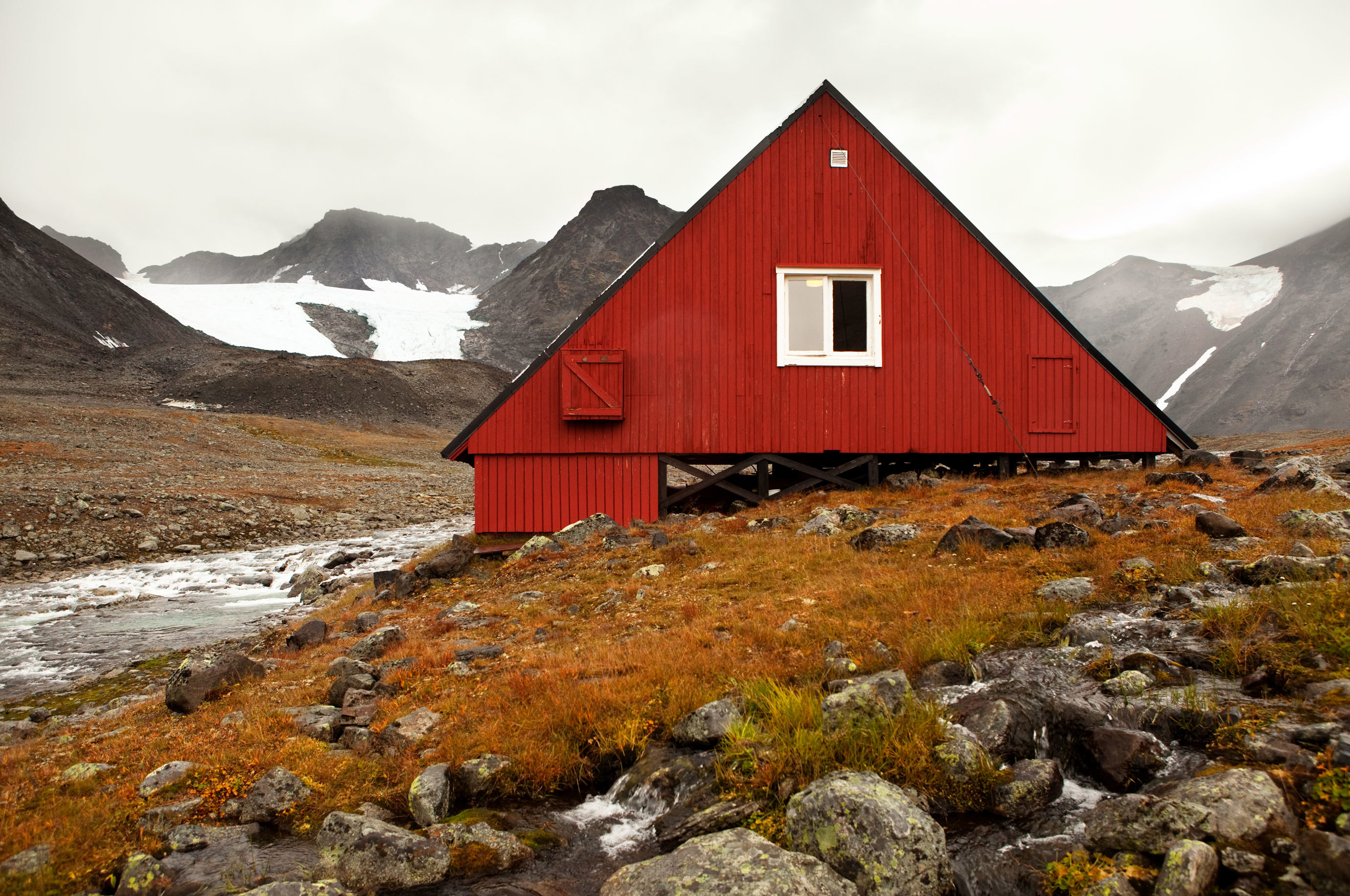 En röd byggnad omgiven av stenar och högfjäll med snöfläckat och glaciärer.