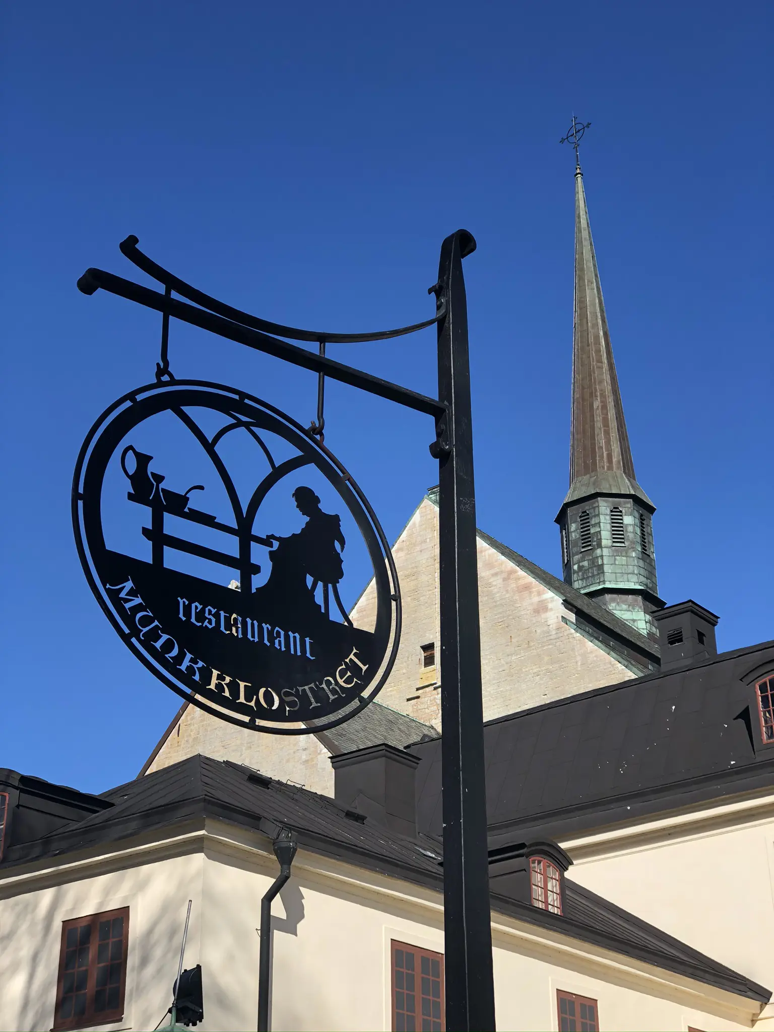 Munkklostret i Vadstena. Smides-skylt för "restaurant Munkklostret". Skylten är rund med en utskuren siluett av en munk som sitter vid ett matbord.