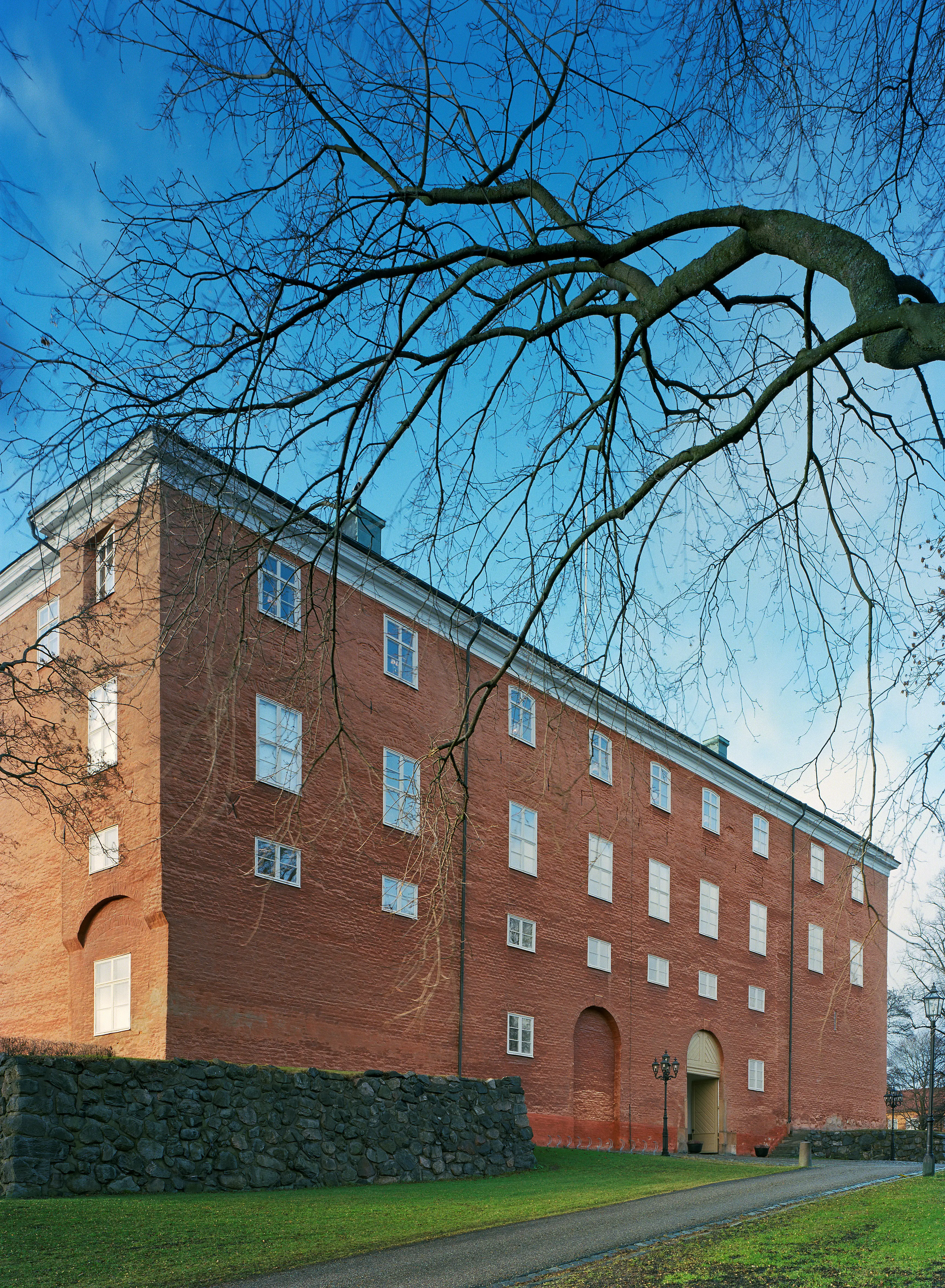 Västerås slott. Byggnaden har fyra våningar. Fasaden är enkel med röd puts på tegel. Tidig vår.