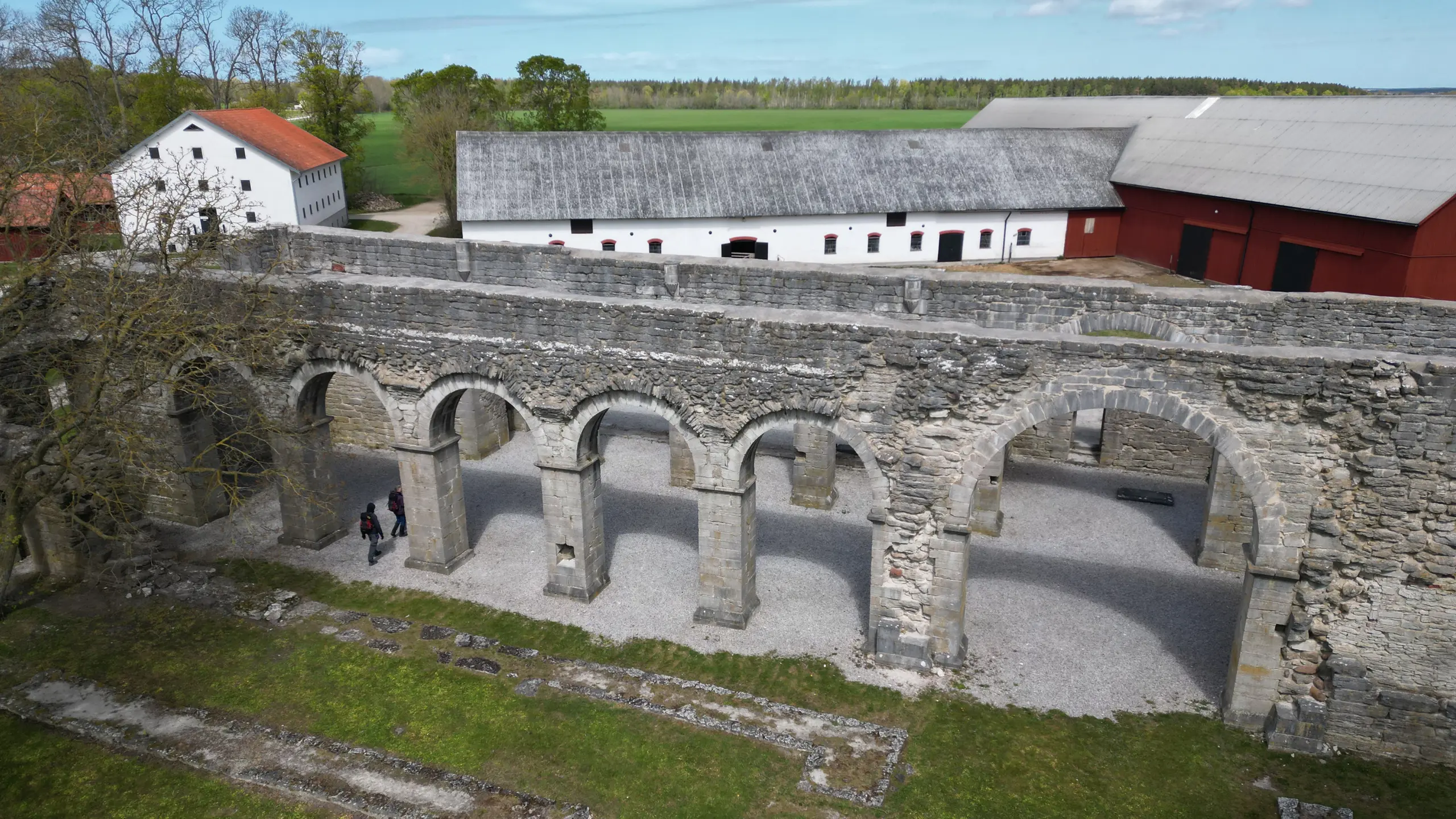 Flygbild av Roma klosterruin. Ett par stenmurar med rundade pelarvalv är intakta. I bakgrunden syns ekonomibyggnader för jordbruk.