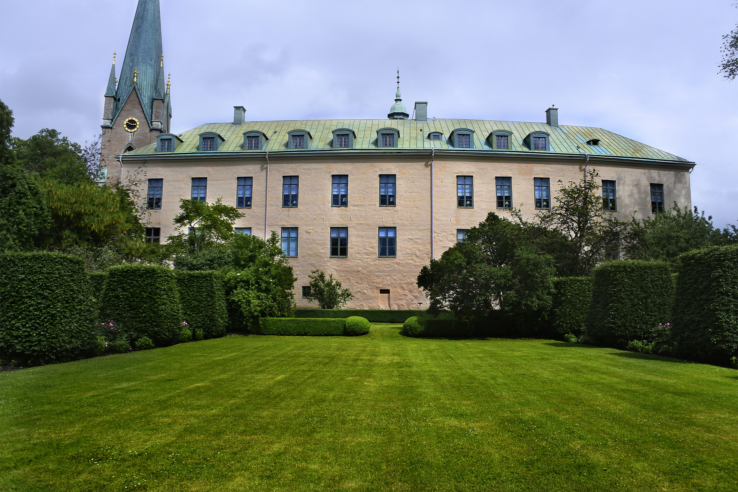Linköpings slott, en avlång byggnad med grönaktigt tak och ett torn till vänster. I förgrunden en välklippt gräsmatta och buskar.