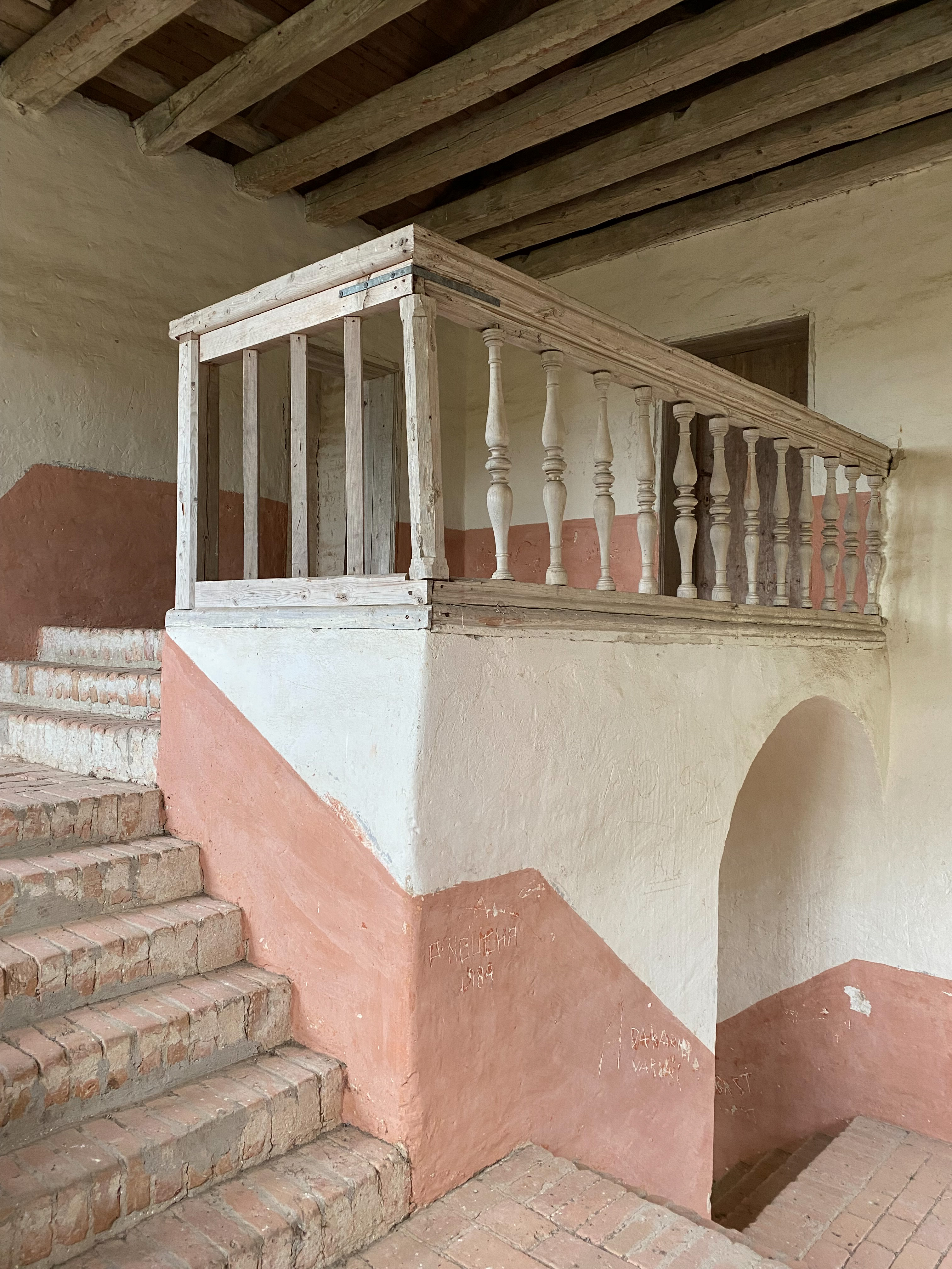 Interiör med putsade väggar, rosa undertill och vita övertill. Ett räcke med svarvade delar i omålat trä avslutar en tegeltrappa...