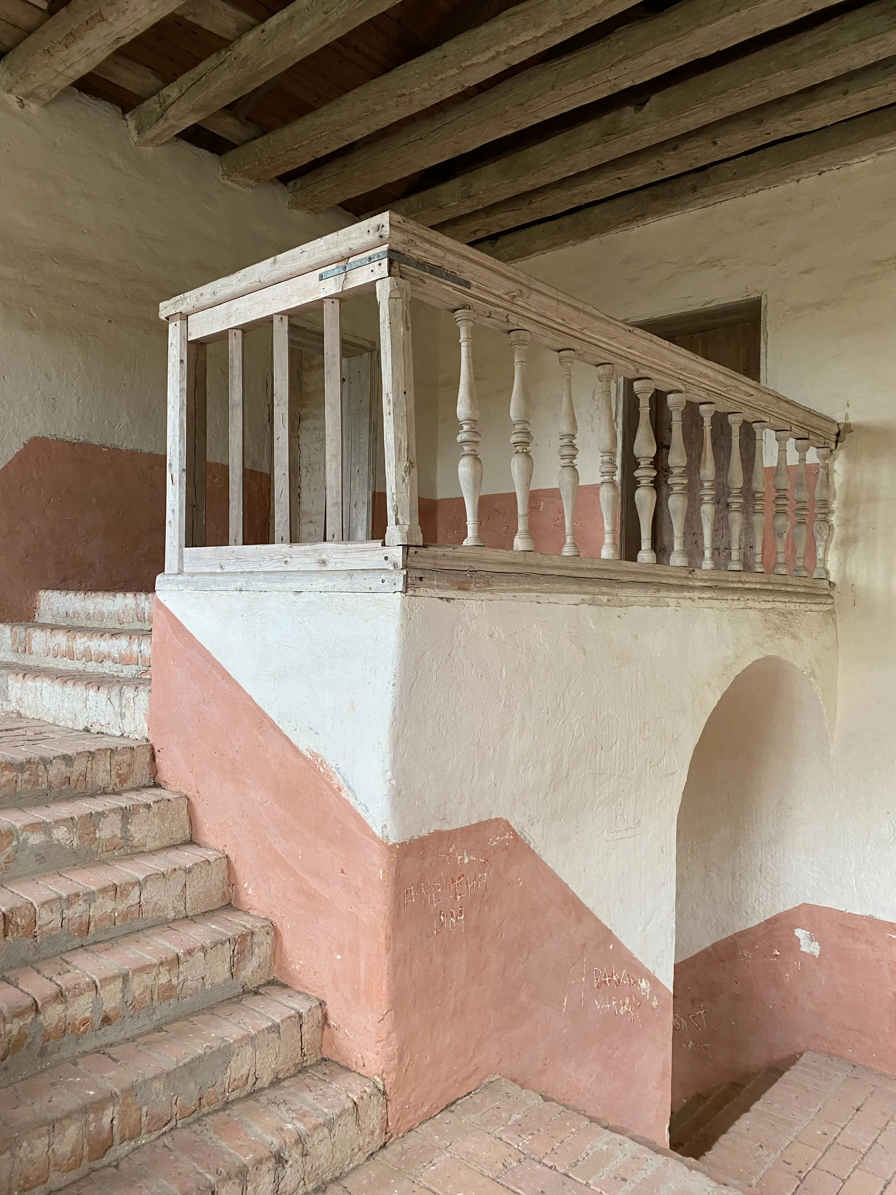 Interiör med putsade väggar, rosa undertill och vita övertill. Ett räcke med svarvade delar i omålat trä avslutar en tegeltrappa...