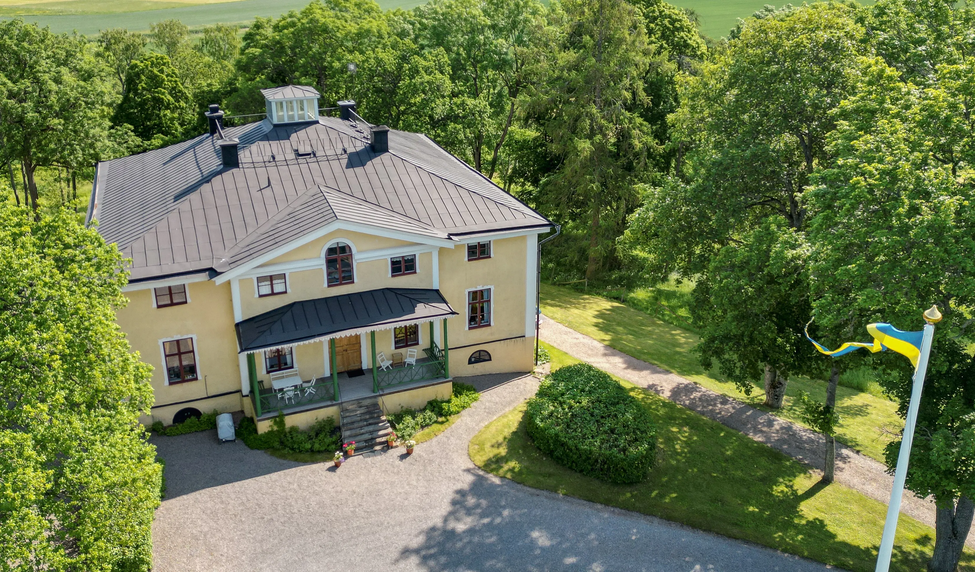 Landsberga. Drönarfoto av stora bostadshuset, en gulputsad villa med entré-veranda och lanternin på taket.