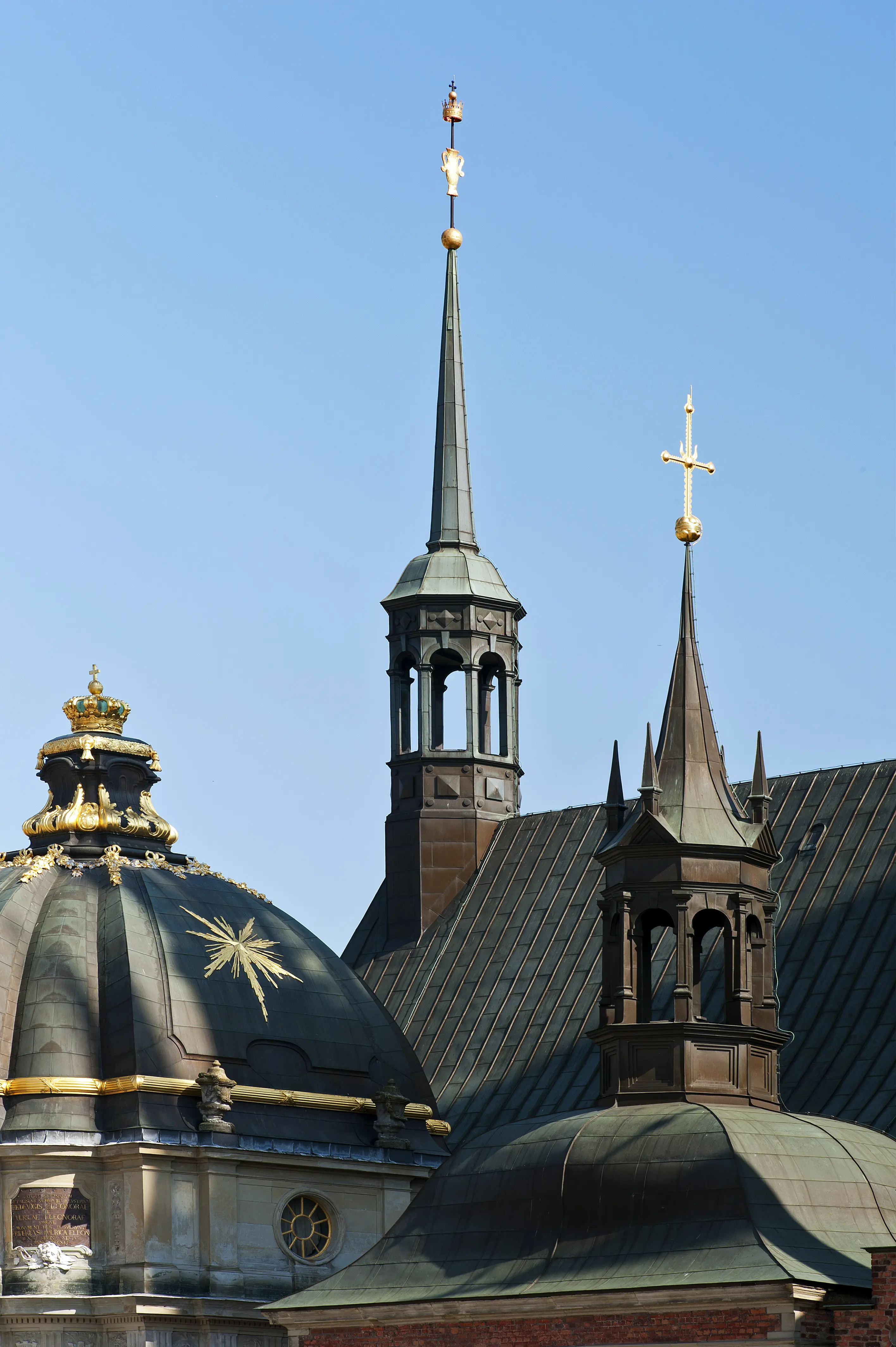 Ett snett mörkt plåttak med två spetsiga kyrktorn och en kupol med svart tak och gulddetaljer. Himlen i bakgrunden är klarblå.