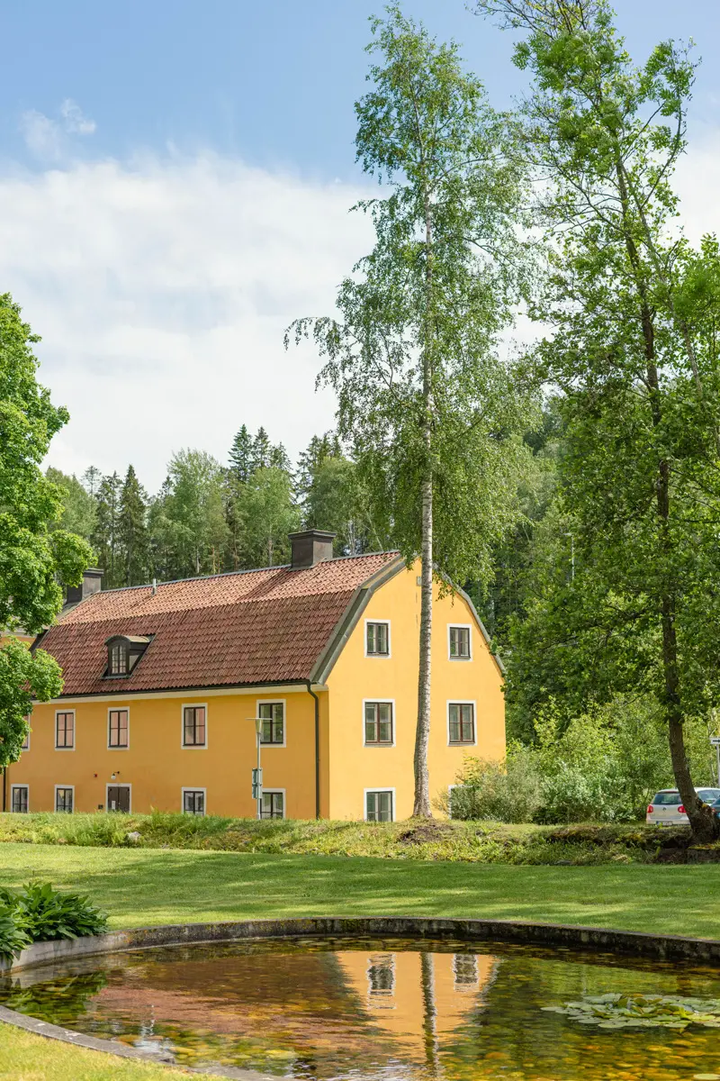 Gulputsat hus med tak av röda tegelpannor. Huset speglas i en rund damm med näckrosor.