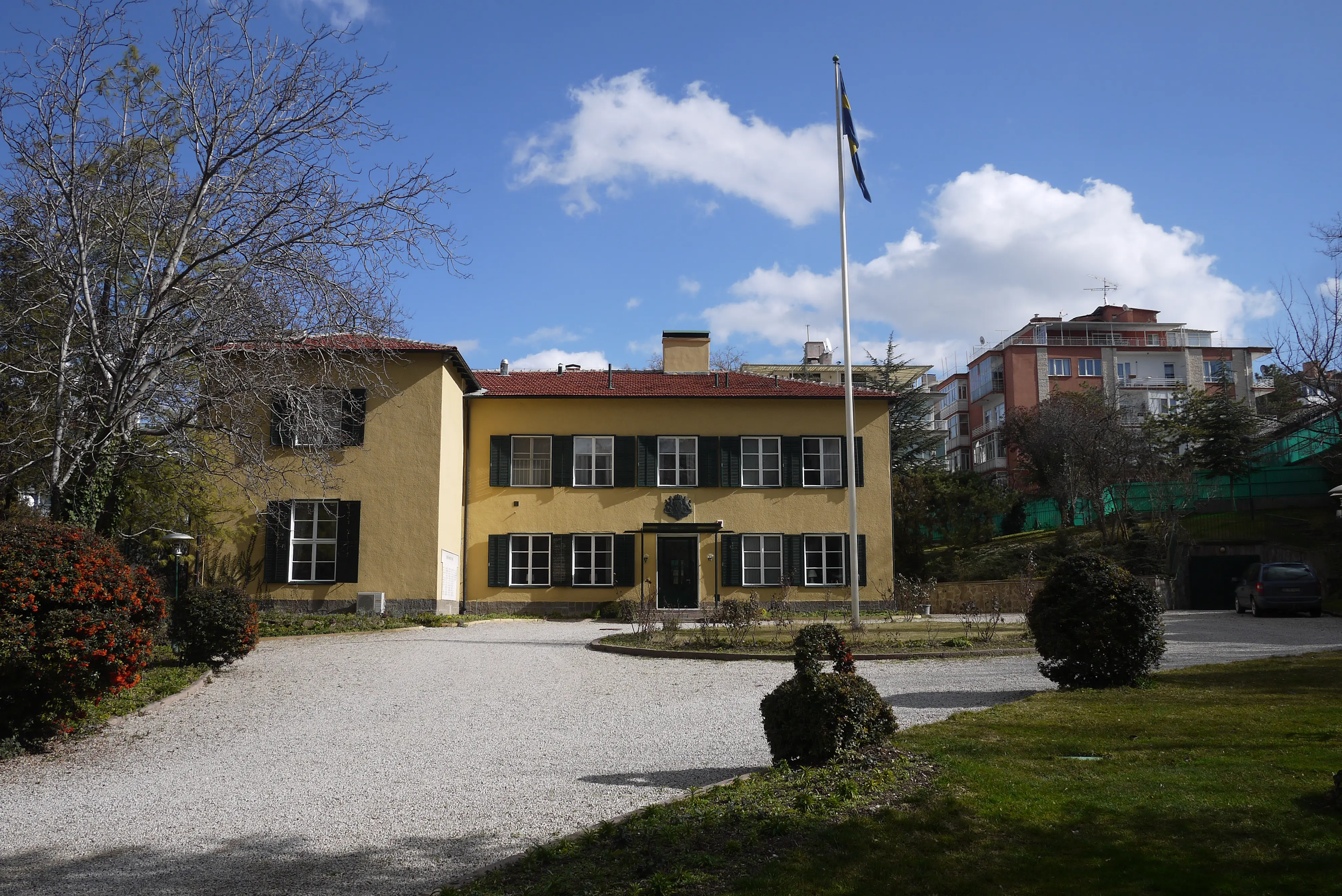 Ambassaden i Ankara. En gulputsad byggnad i två våningar. En bred grusväg leder fram till huset, och kantas av planteringar och träd.