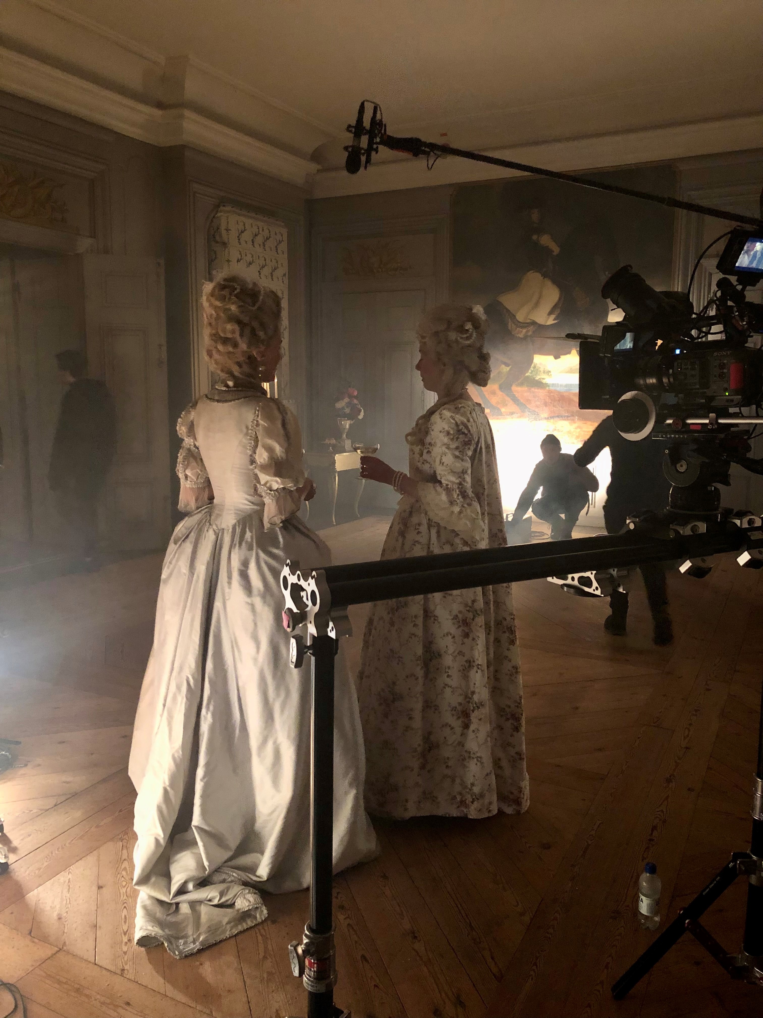 Filminspelning i en salong på ett slott. Två personer i historisk dräkt står bland filmutrustning.