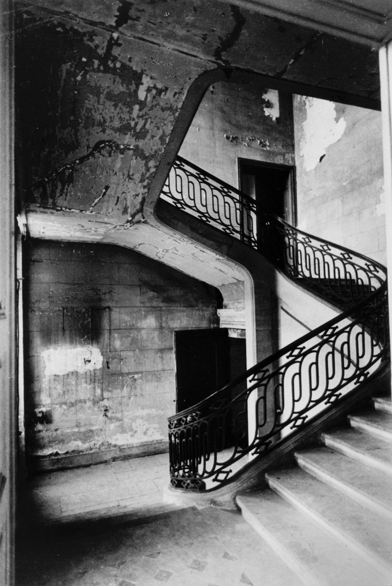 Svenska institutet i Paris. Svartvit bild av entré-hallen med sin stora, svängda stentrappa med smidesräcke. Mycket slitet.