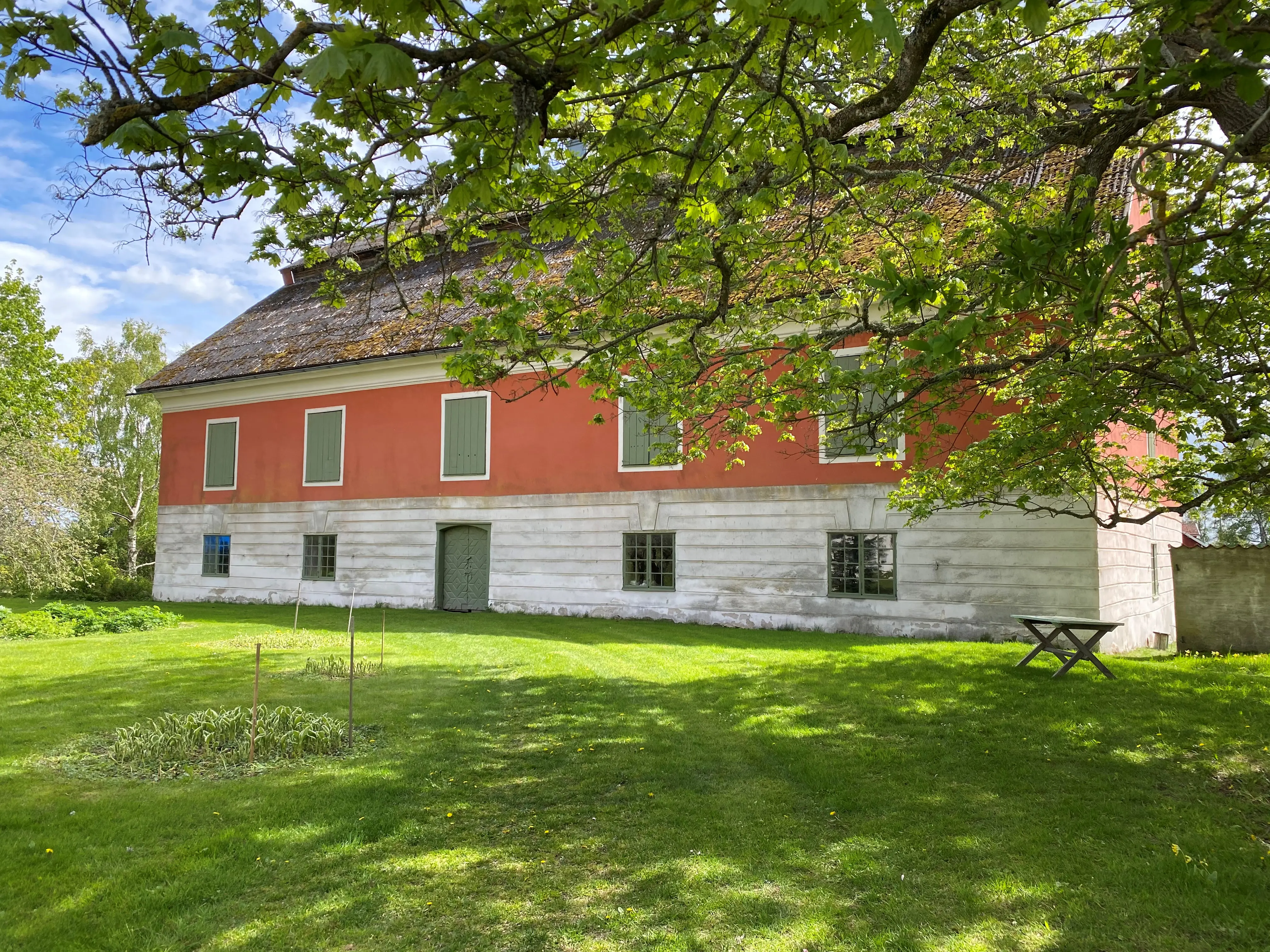 Stora Stenhuset på Tuna kungsgård. Tvåvåningsbyggnad putsad i vitt och rött, med gröna fönsterluckor. Vår, blommor och skir grönska.