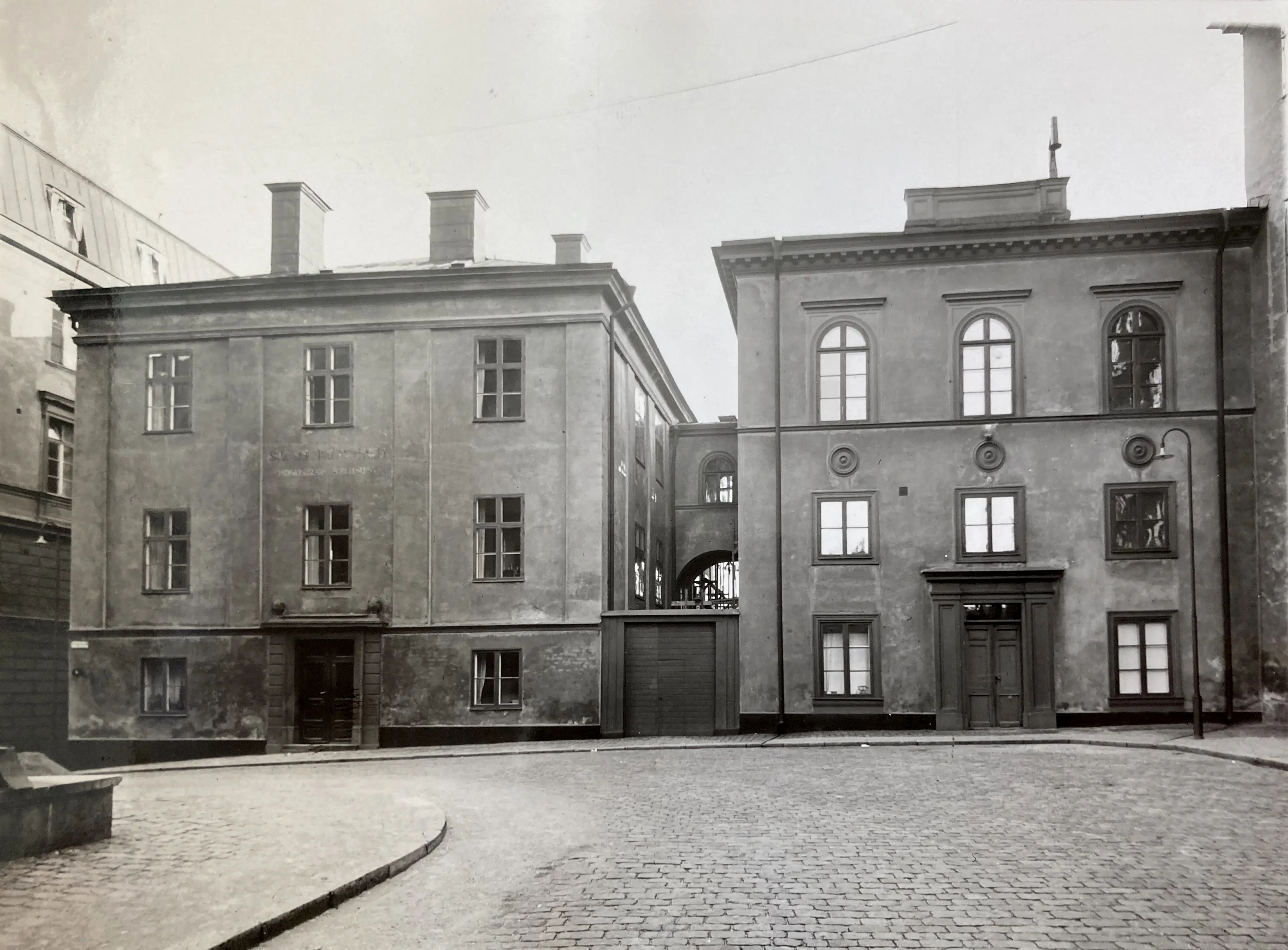 Svartvitt foto på två stenhus bredvid varandra. I förgrunden en kullerstensgata.