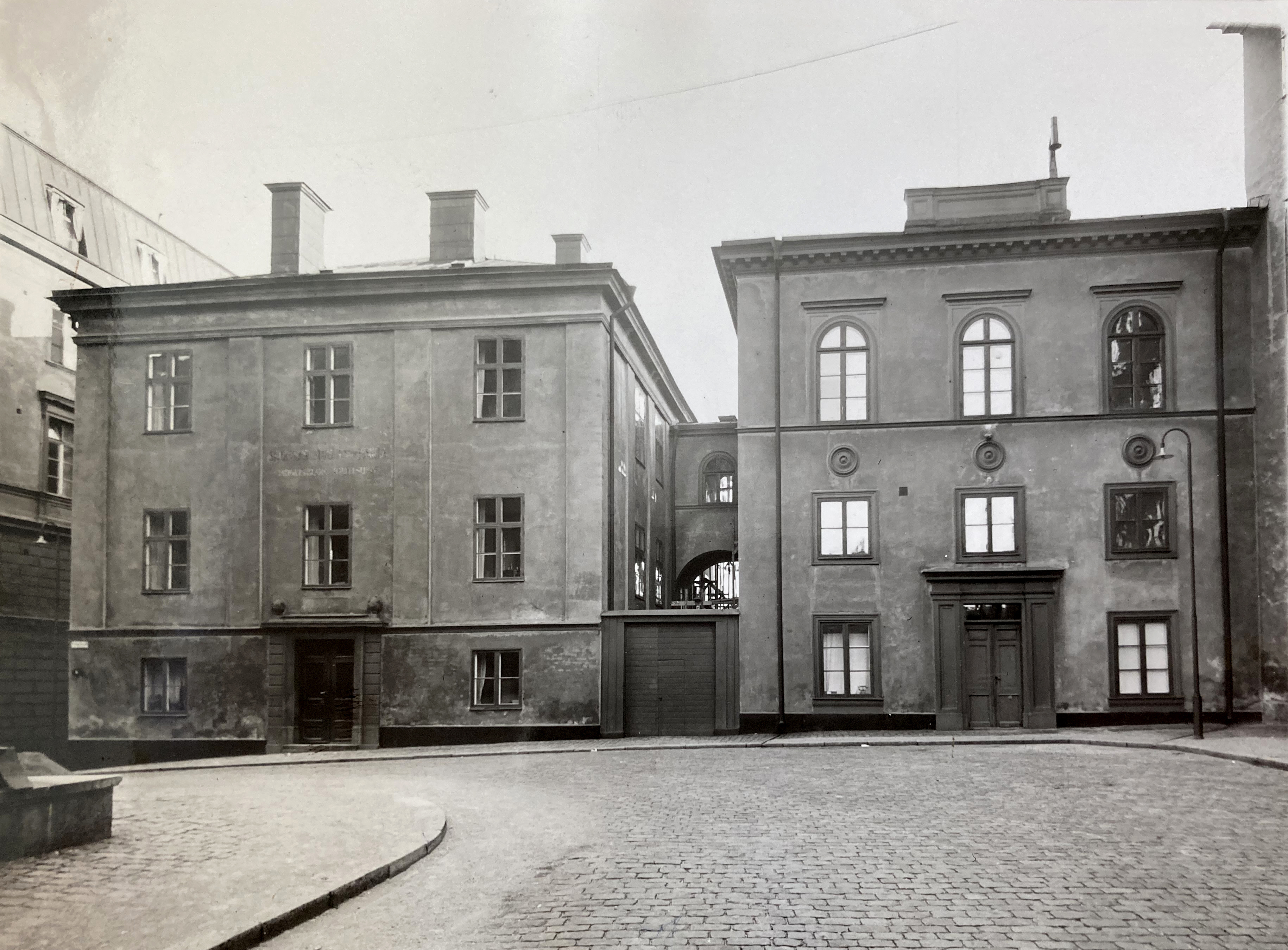 Svartvitt foto på två stenhus bredvid varandra. I förgrunden en kullerstensgata.