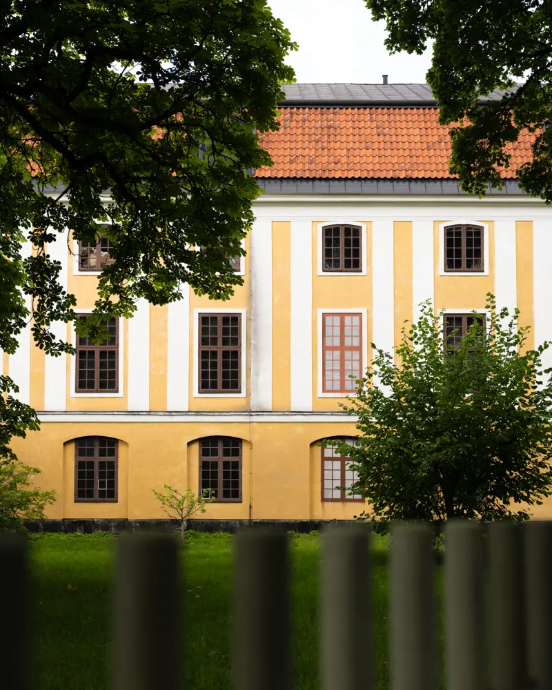 Confidencens fasad i gul och vit puts. Taket har röda tegelpannor. I förgrunden syns ett staket och lövverket från flera träd.