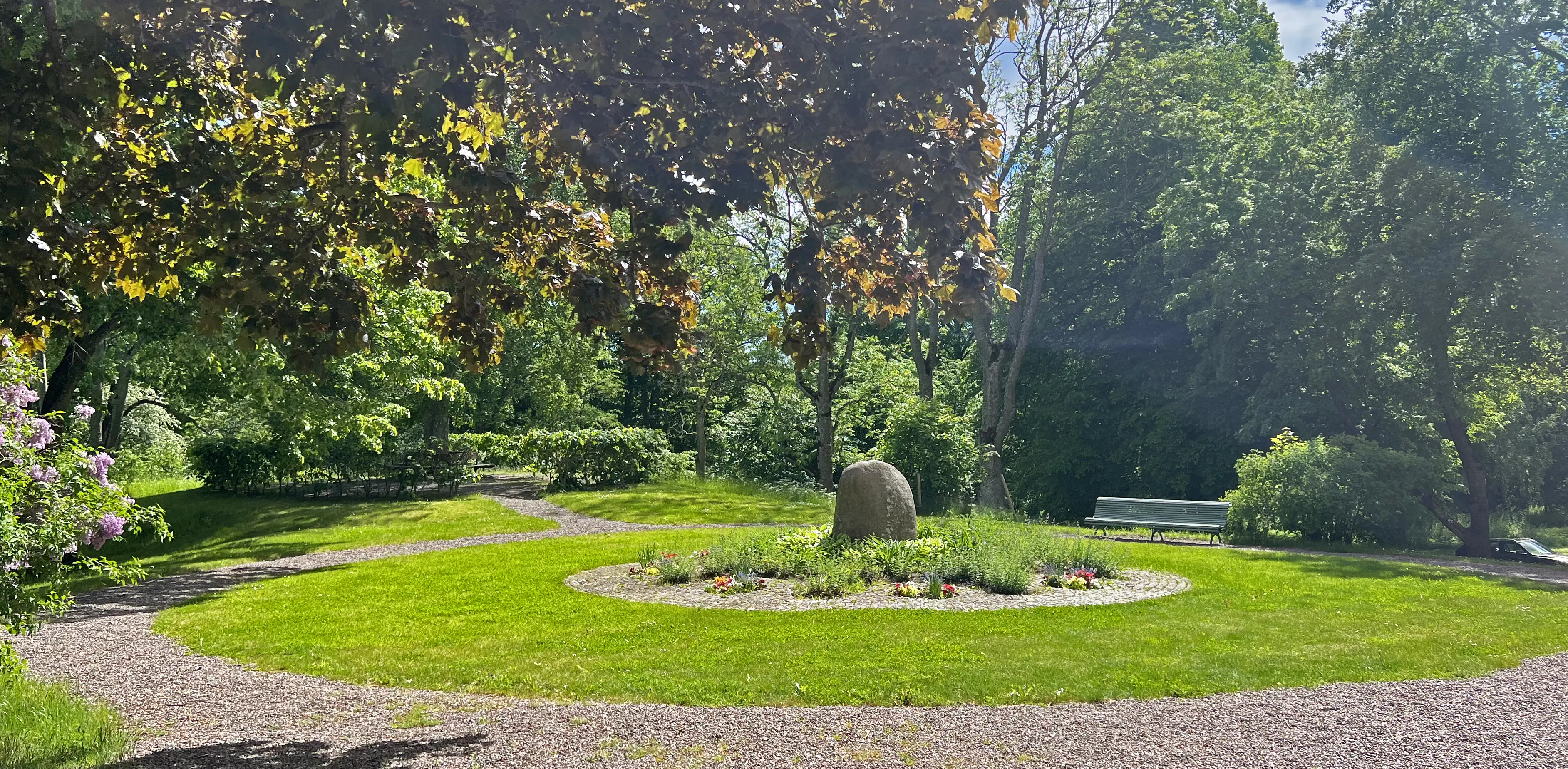 Bogesunds slottsträdgård. Brahestenen, inramad av blomsterplanteringar, gräsytor, grusgångar och sittbänkar. Sommar och grönska.