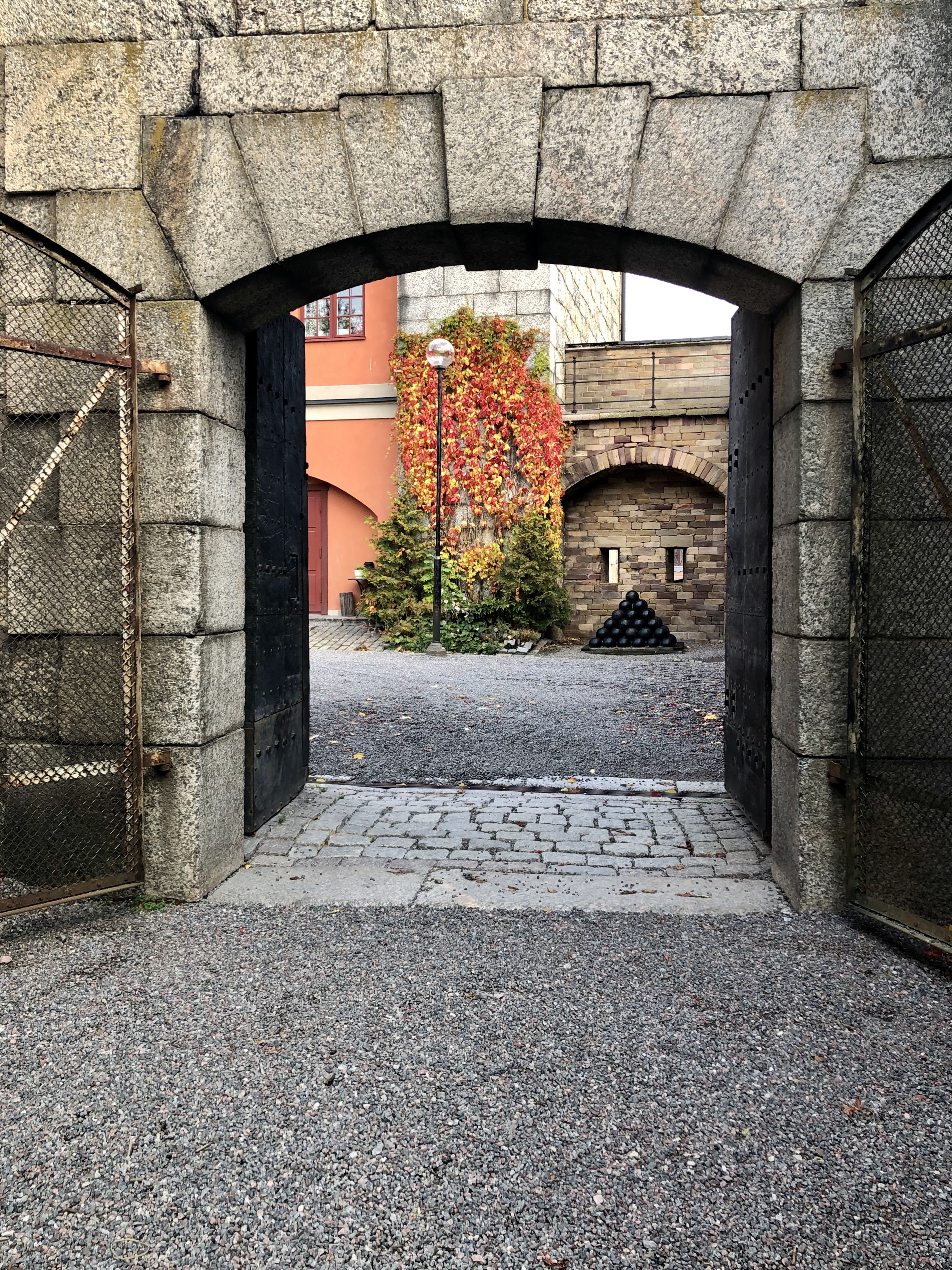 Portvalv i stenmuren kring Vaxholms kastell. De öppna portarna är av järn. Genom öppningen syns en byggnad med höstglödande murgröna.