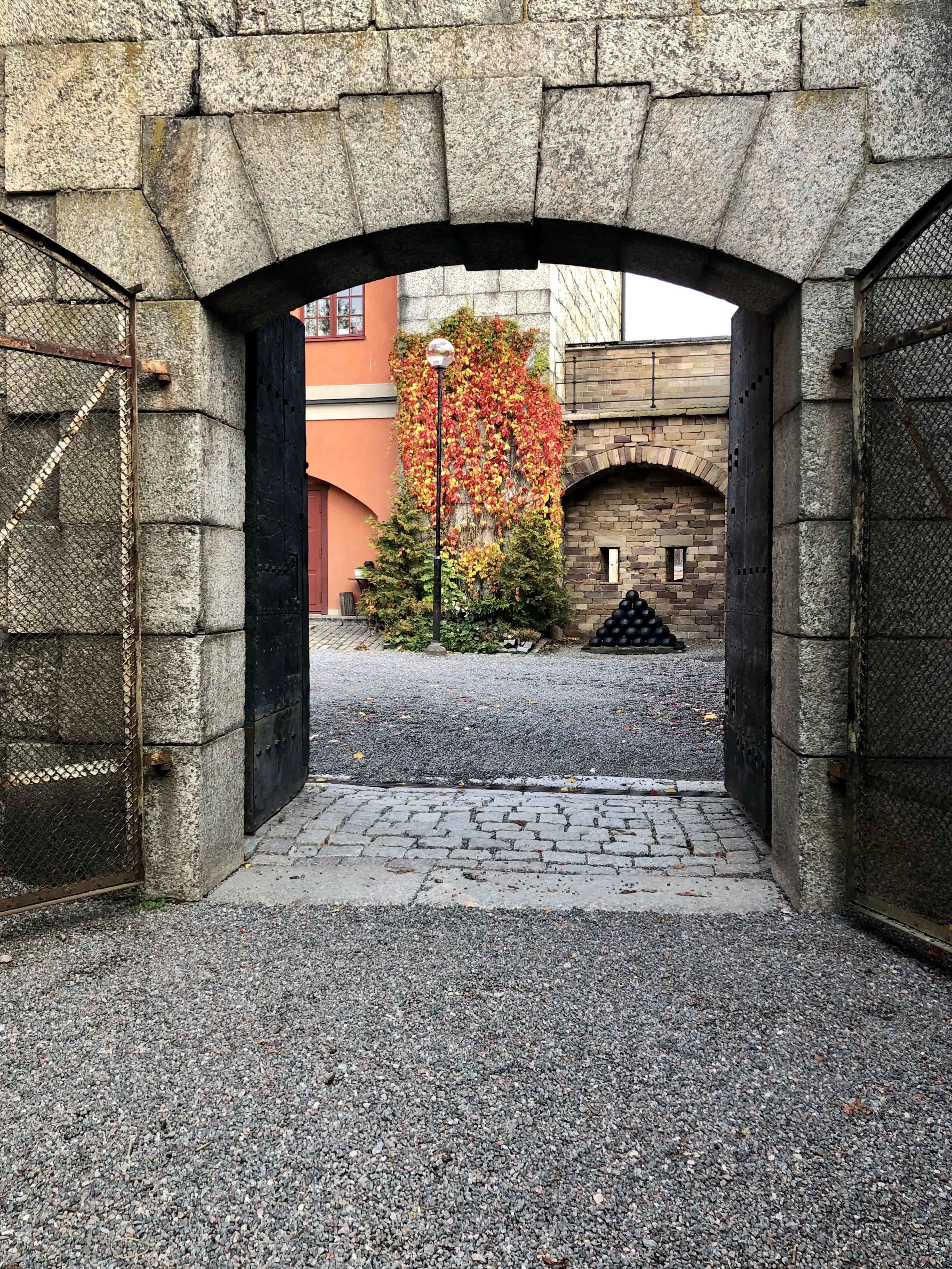 Portvalv i stenmuren kring Vaxholms kastell. De öppna portarna är av järn. Genom öppningen syns en byggnad med höstglödande murgröna.