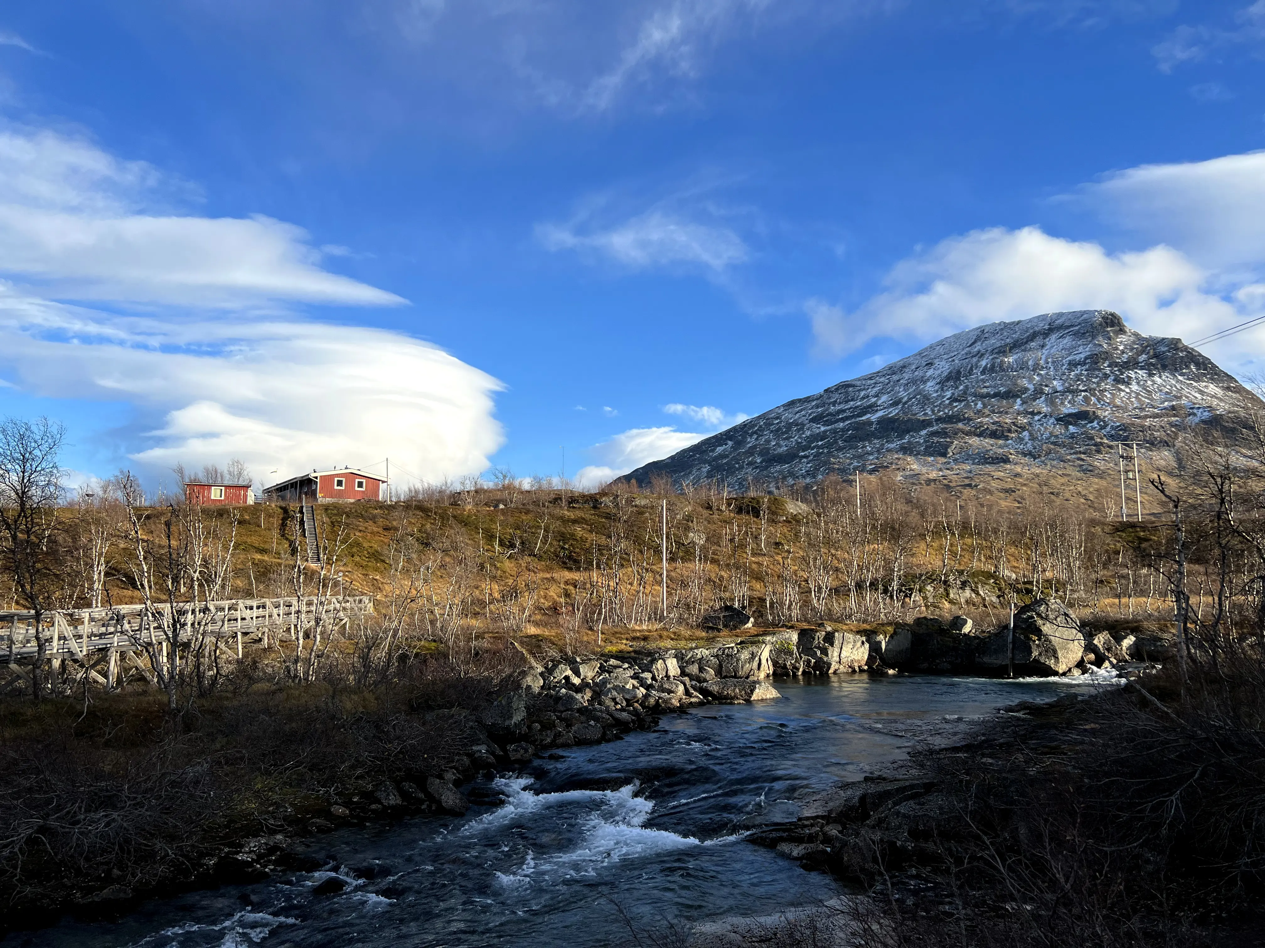 Ett fjällandskap med snöbeklädda toppar tornar upp sig bakom två röda hus med vita knutar. En spång över ett vattendrag leder upp till husen.
