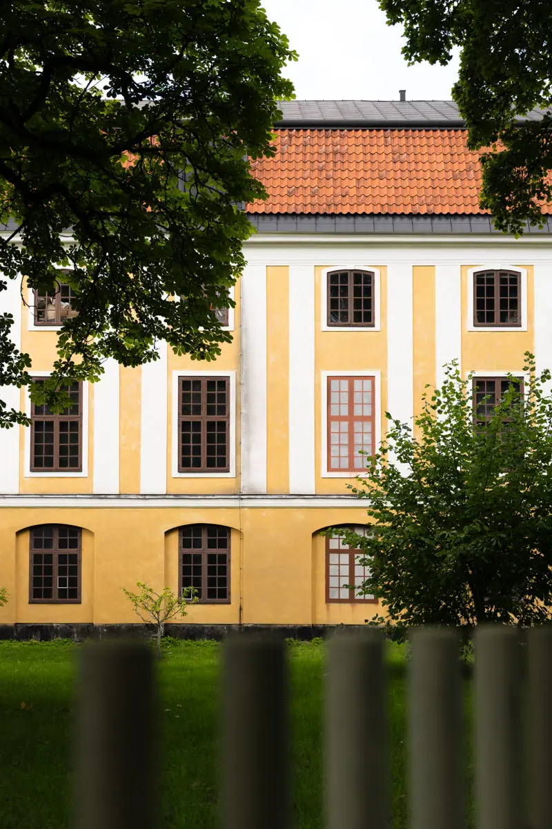 Confidencens fasad i gul och vit puts. Taket har röda tegelpannor. I förgrunden syns ett staket och lövverket från flera träd.