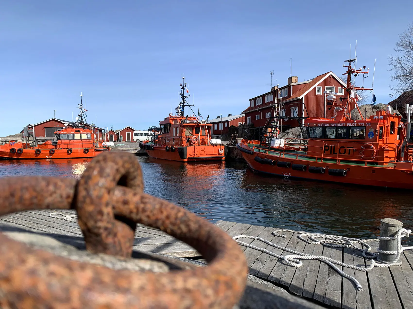 Hamnen på Öja. Röda trähus och röda lotsbåtar. Längs träkajerna sitter olika förtöjnings-anordningar, som rep och grova järnringar.