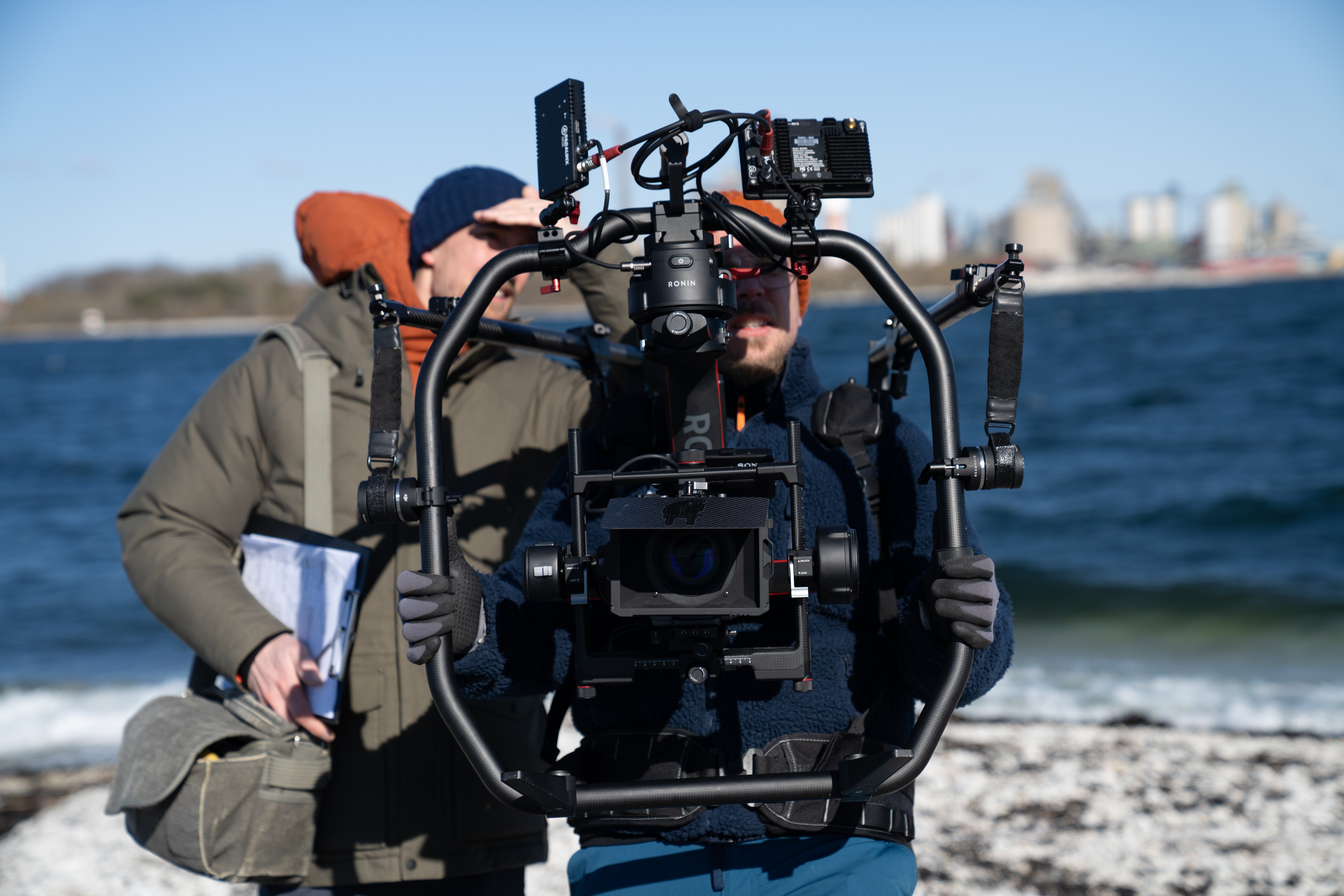 Två personer står med filmutrustning på en stenig strand. 