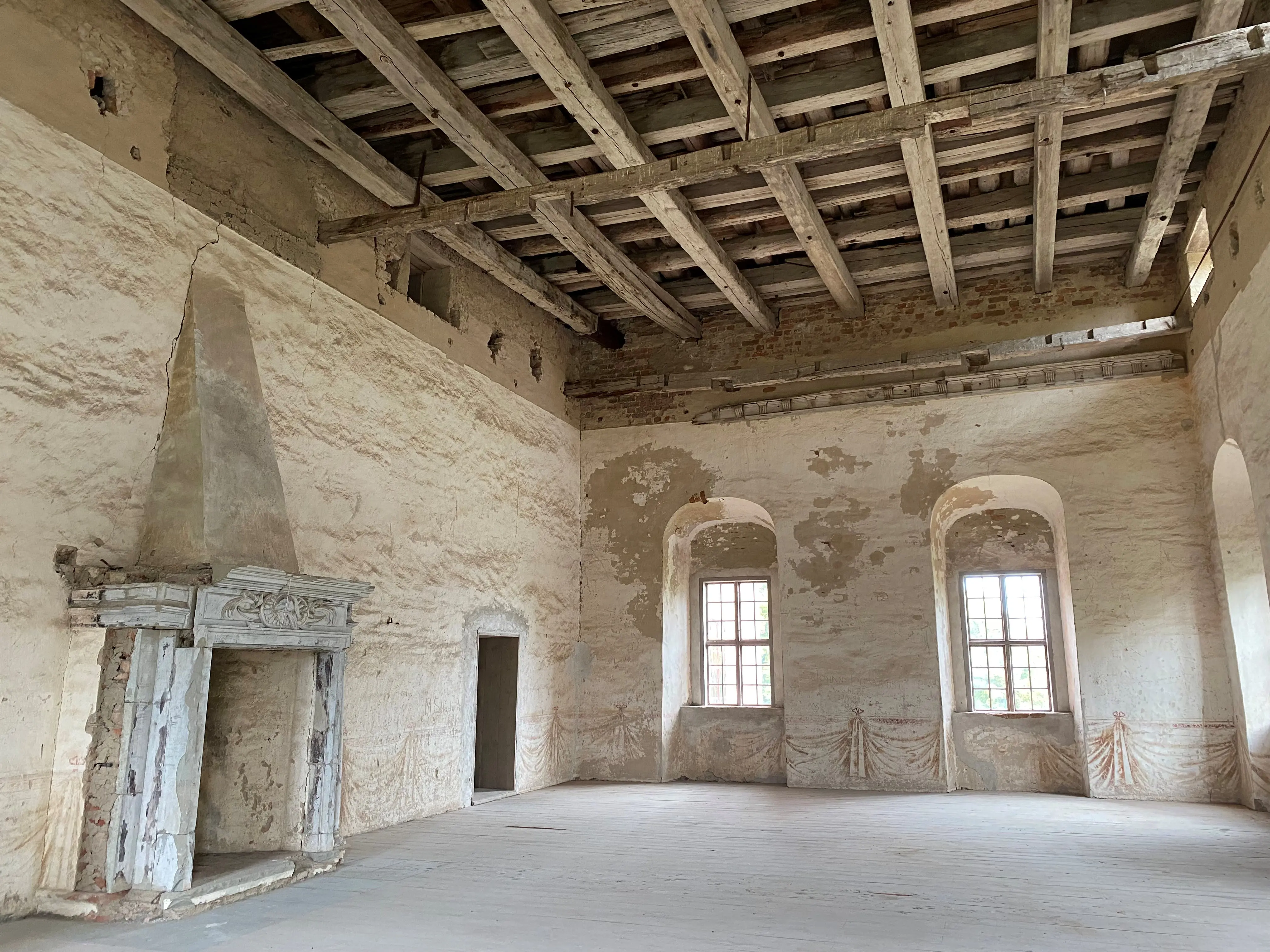 Interiör gammalt rum med vitpusade väggar en stor öppen spis med dekorerad omfattning samt stor fönster. I taket syns bjälkarna.