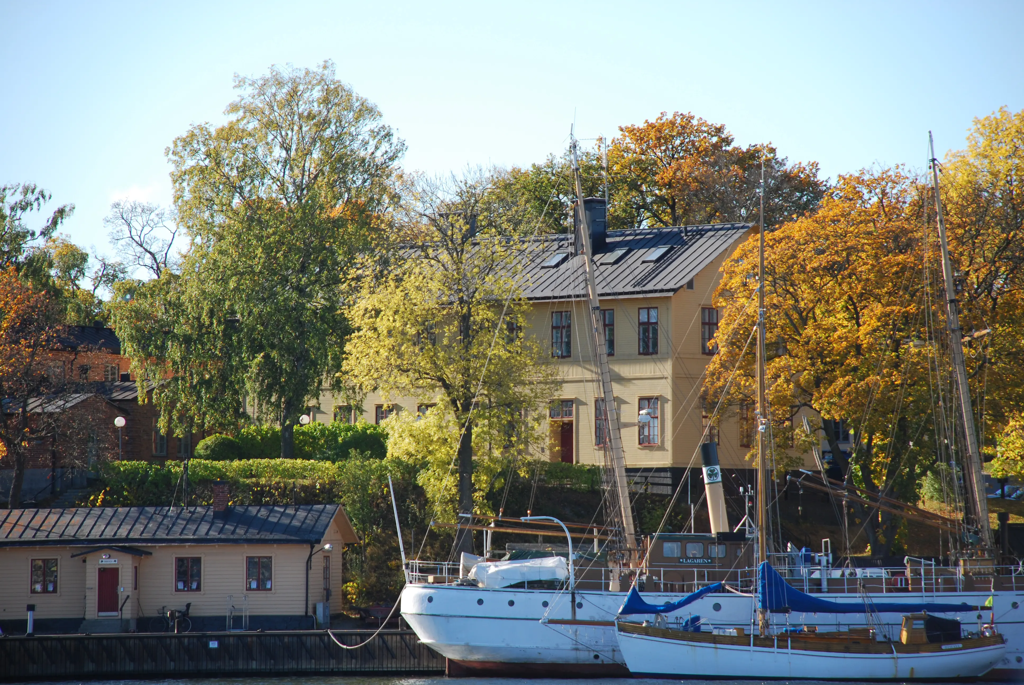 Sjöreservens kasern på Skeppsholmen. Gul träbyggnad omgiven av höstgula träd. I förgrunden kaj och båtar.