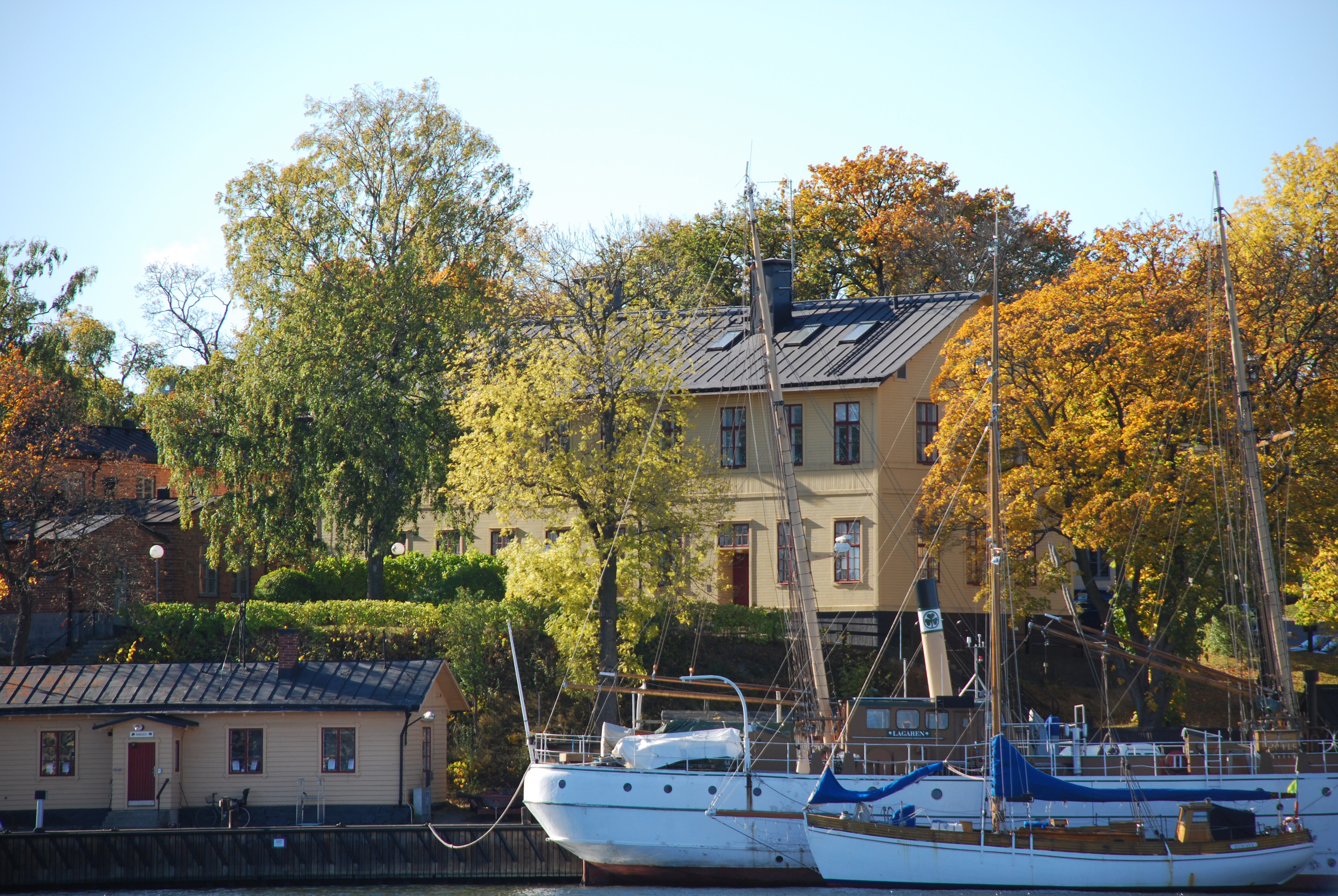 Sjöreservens kasern på Skeppsholmen. Gul träbyggnad omgiven av höstgula träd. I förgrunden kaj och båtar.